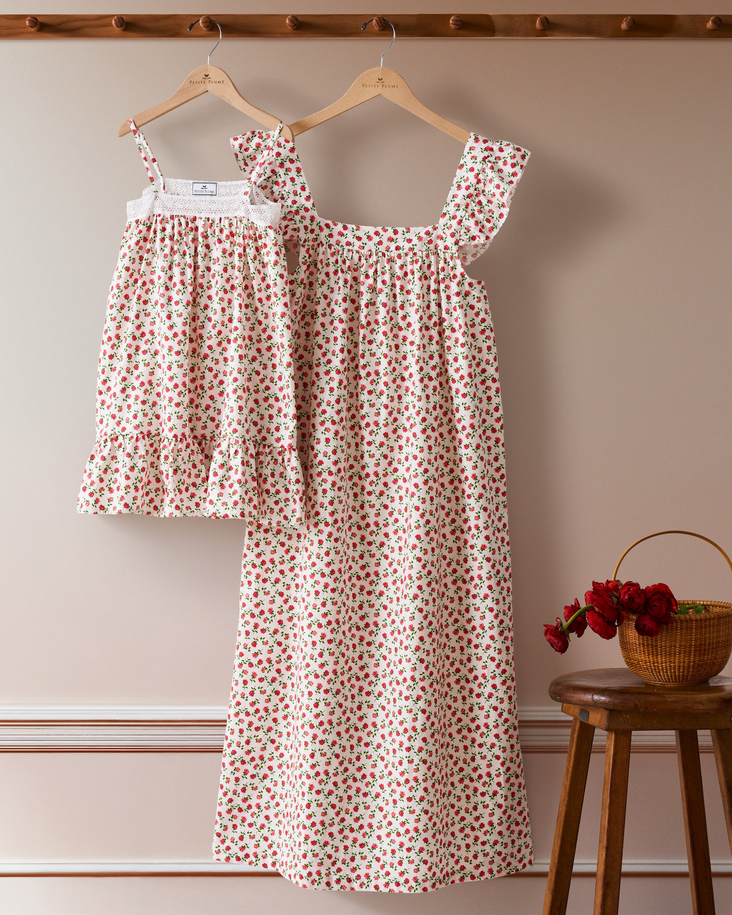Two matching Petite Plume Girls Lily Nightgowns in Petite Petals, with red floral patterns, hang on wooden hangers against a beige wall. A wicker basket with red flowers rests on a stool to the right. The larger gown is made from yarn-dyed cotton and features ruffled shoulder straps.