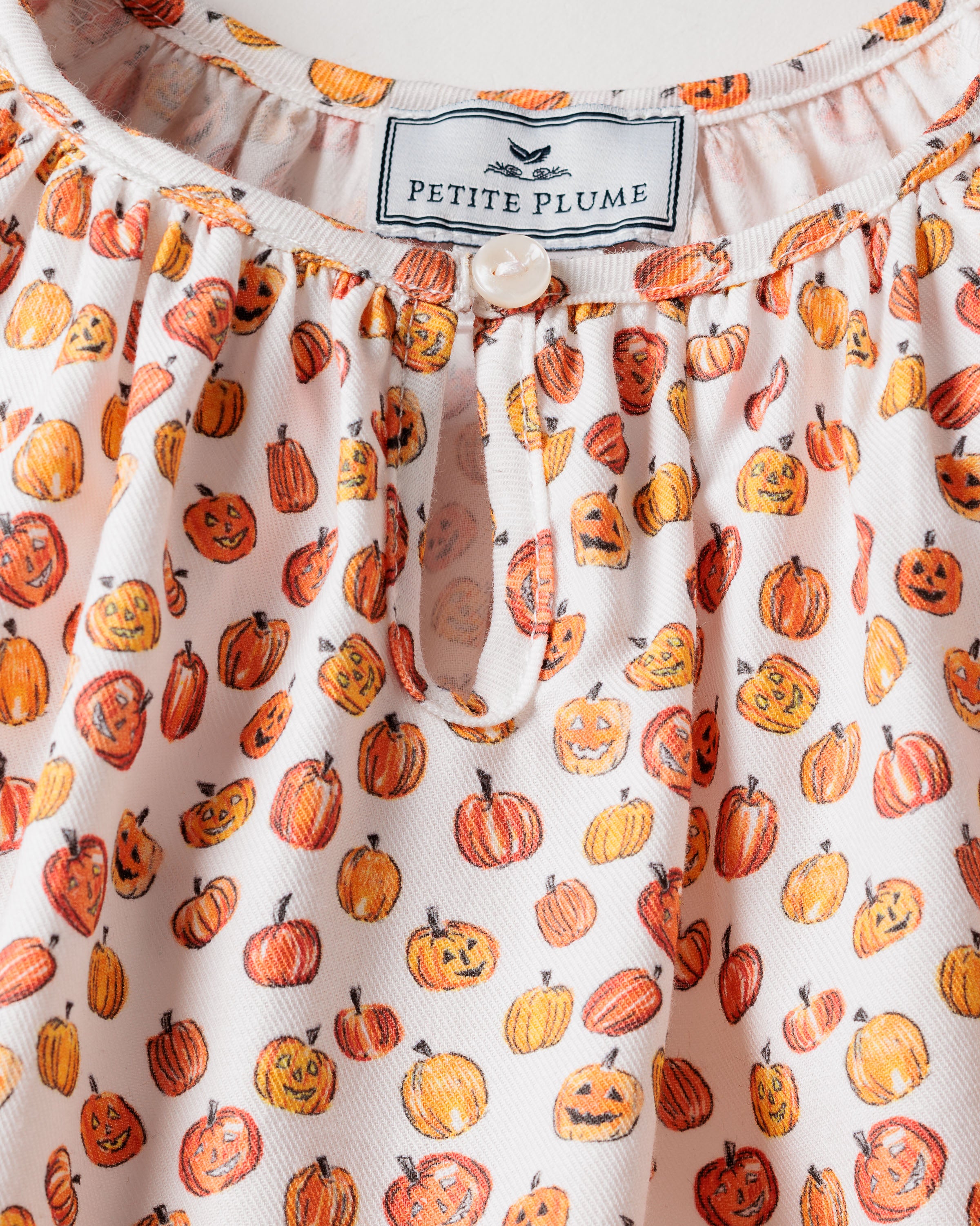 The Girls Delphine Nightgown in Pumpkin Patch by Petite Plume features small orange jack-o-lanterns with various expressions on childrens sleepwear. The garment, made from delightful chemical-free fabric, has the Petite Plume label near the neck area.