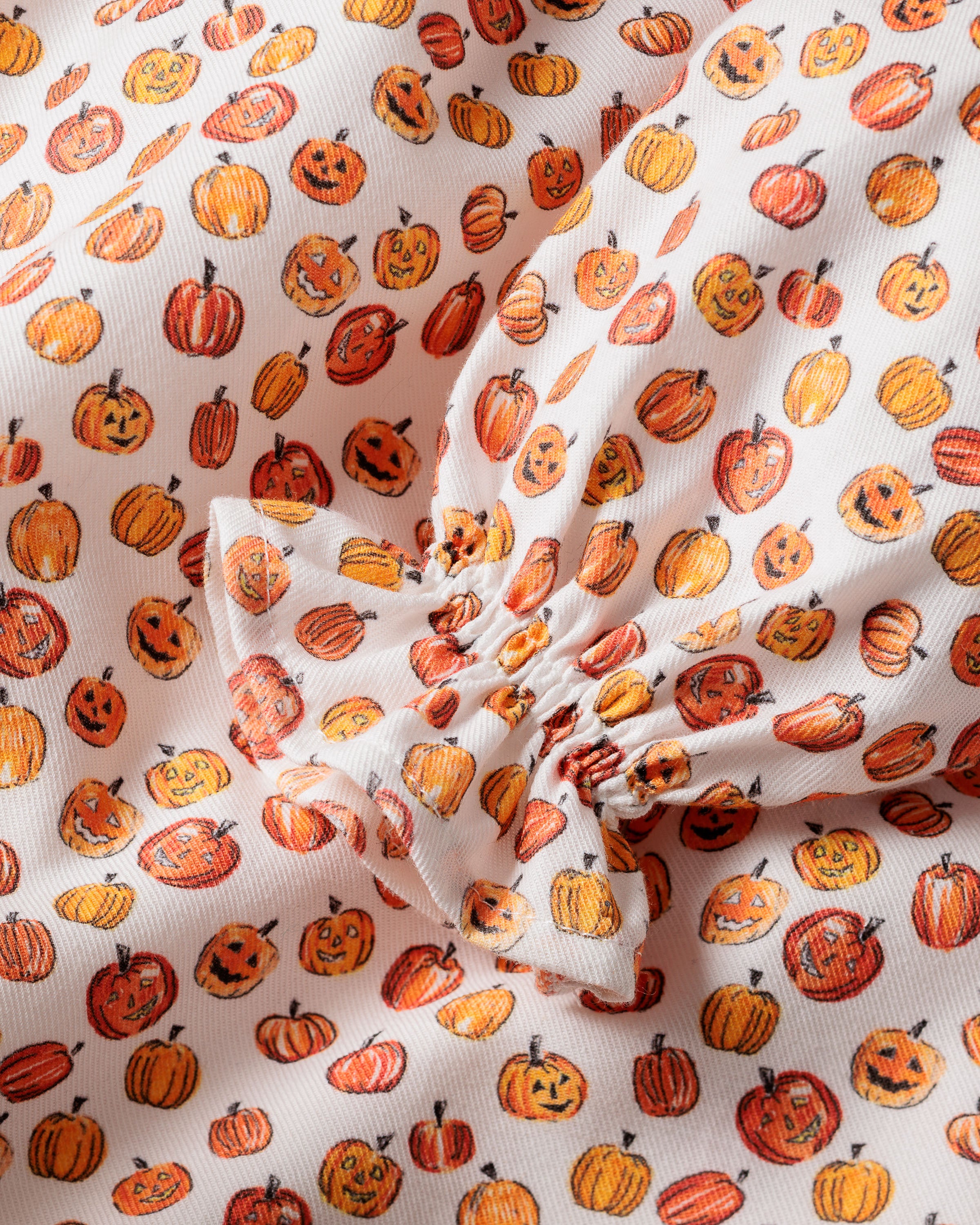 The Girls Delphine Nightgown in Pumpkin Patch by Petite Plume features a playful print with small orange jack-o-lanterns and pumpkins on a white background, with gathered fabric in the center, adding texture ideal for childrens sleepwear.