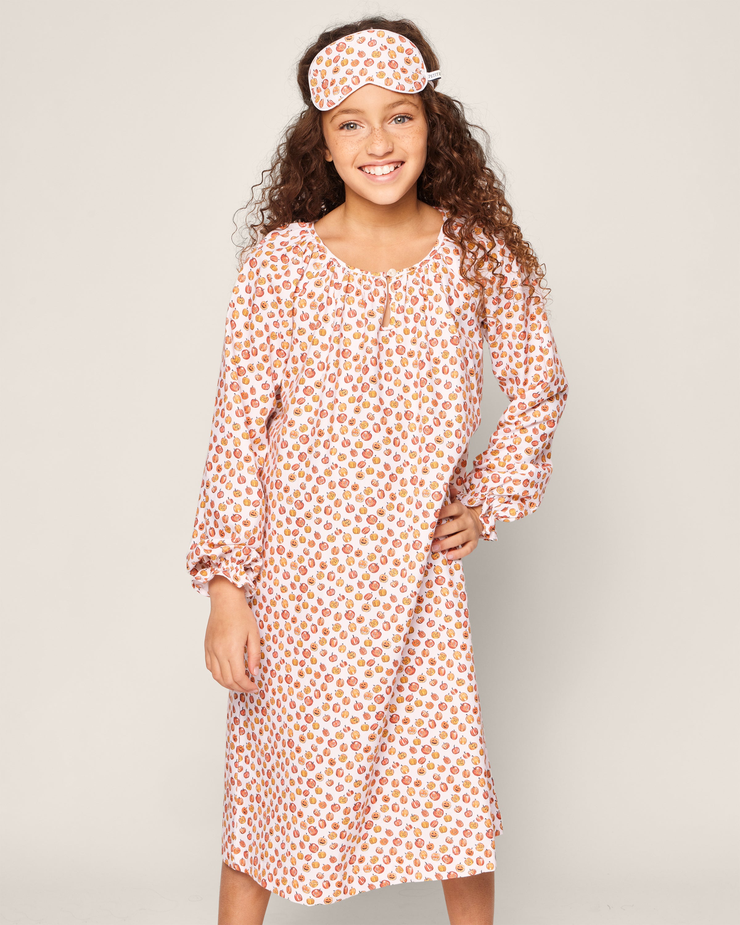 A young girl with curly hair smiles in a Petite Plume Girls Delphine Nightgown in Pumpkin Patch. Made from chemical-free fabric, its red floral pattern and matching sleep mask display charming kids’ print against a plain light background.