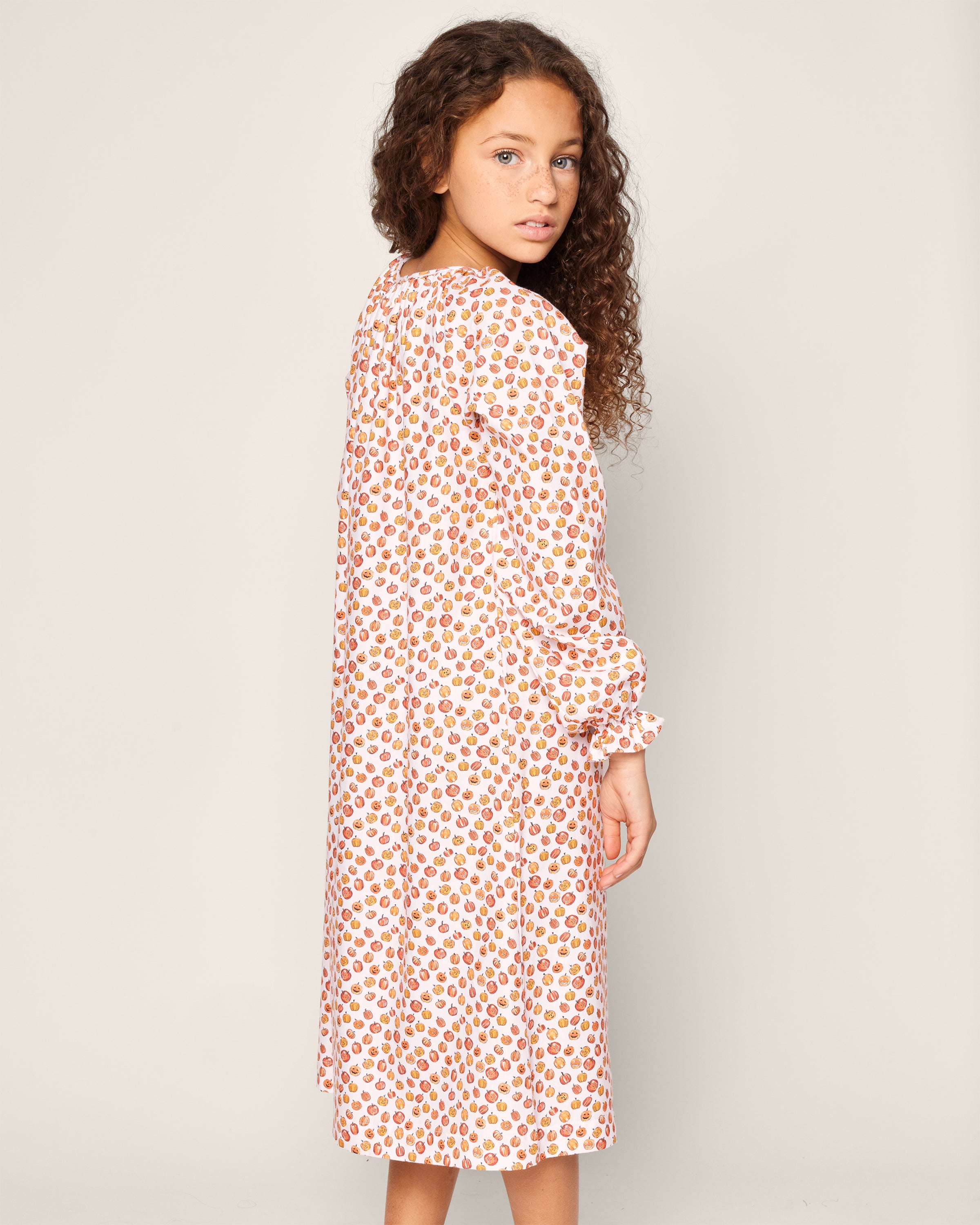 A young girl with long, curly hair is wearing the Petite Plume Girls Delphine Nightgown in Pumpkin Patch, featuring a vibrant red and orange floral design. She stands sideways against a plain background, looking over her shoulder.
