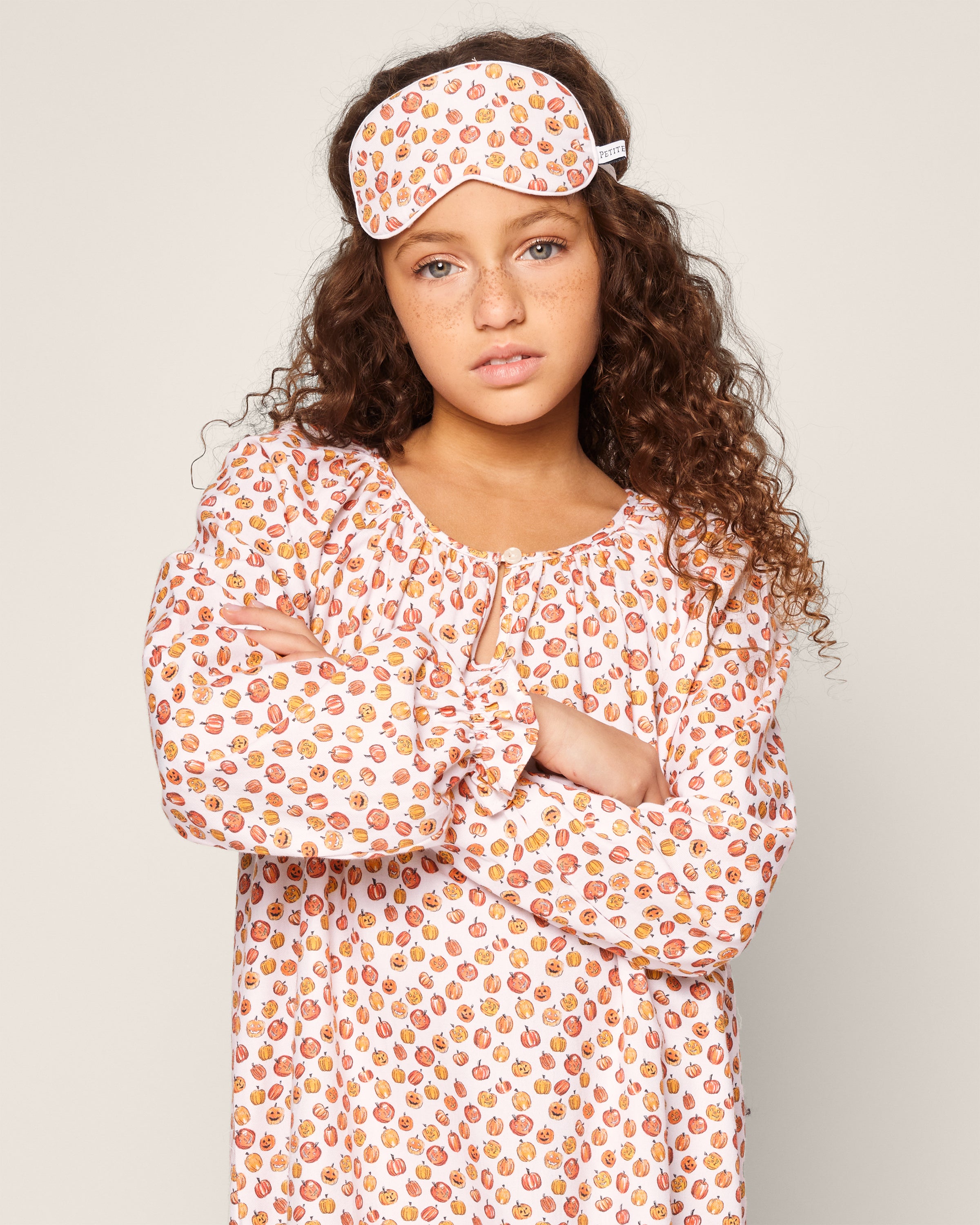 A girl with curly hair stands with crossed arms against a plain background wearing the Petite Plume Girls Delphine Nightgown in Pumpkin Patch, complete with a vibrant kids print of colorful fruits and a matching sleep mask.
