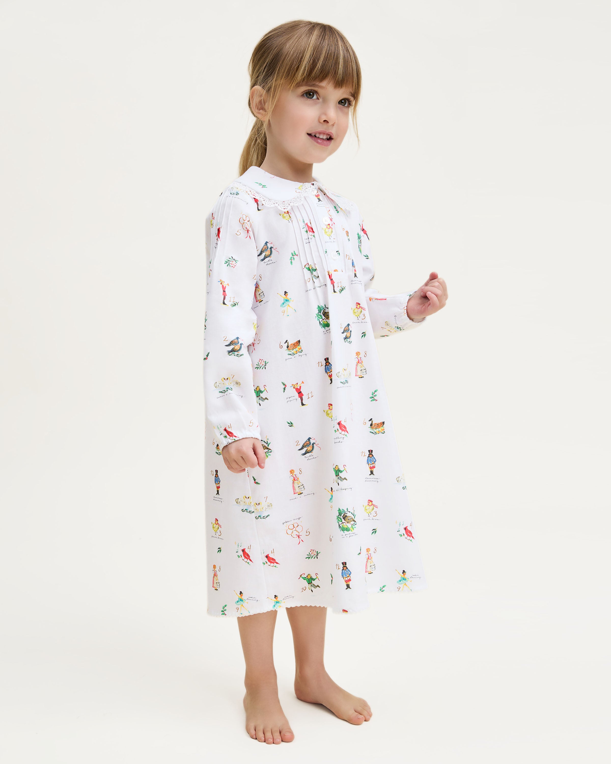 The Girl's Twill Lola Nightgown in 12 Days of Christmas is a white long-sleeve children’s dress with a Peter Pan collar and a playful pattern of colorful people, animals, and plants scattered across the fabric.