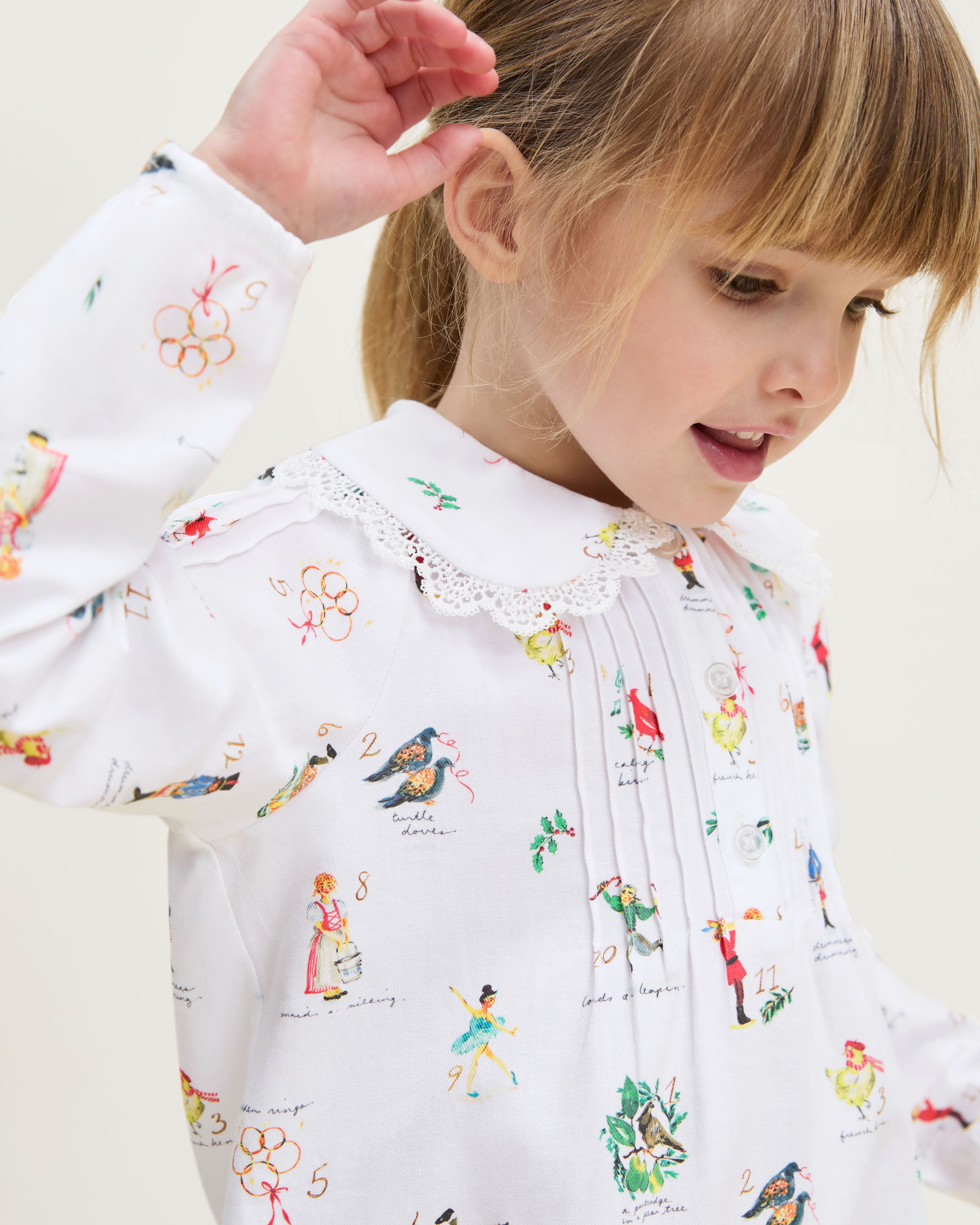 The Girl's Twill Lola Nightgown in 12 Days of Christmas is a white long-sleeve children’s dress with a Peter Pan collar and a playful pattern of colorful people, animals, and plants scattered across the fabric.