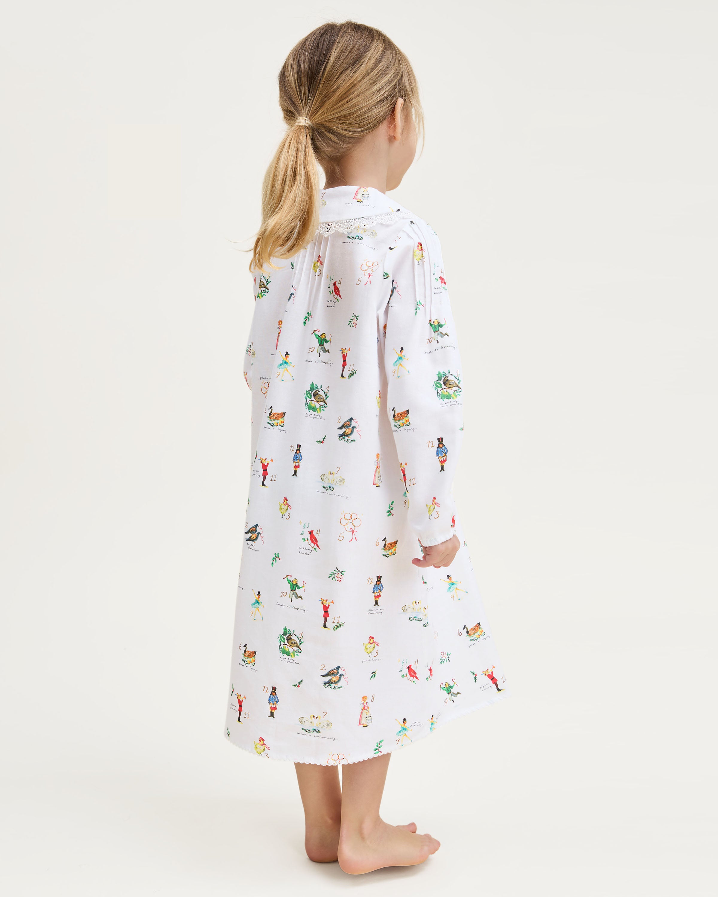 The Girl's Twill Lola Nightgown in 12 Days of Christmas is a white long-sleeve children’s dress with a Peter Pan collar and a playful pattern of colorful people, animals, and plants scattered across the fabric.