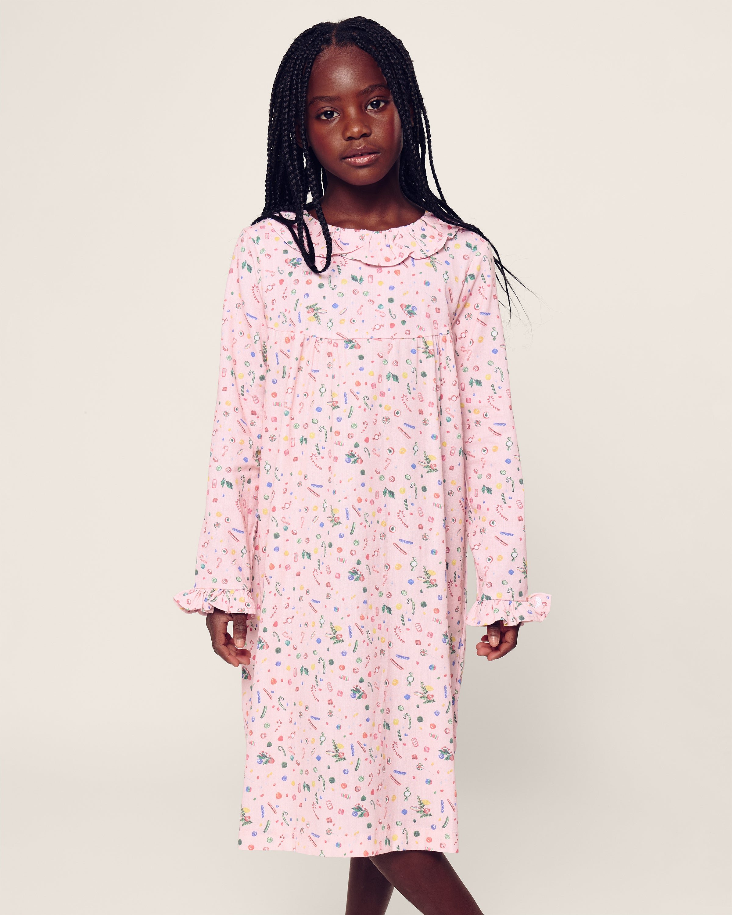 A young girl in Petite Plumes Girls Twill Scarlett Nightgown (Vintage Sweets) stands against a plain background. The flame-retardant sleepwear with long sleeves, ruffled cuffs and neckline showcases small colorful patterns. Her long braided hair complements her calm expression.