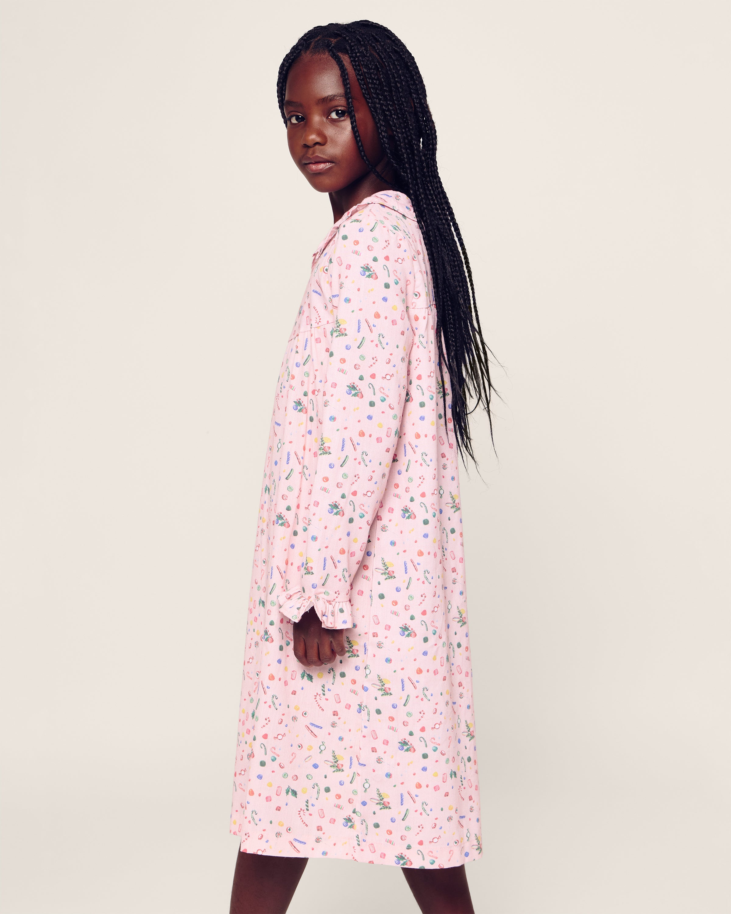 The person is in profile wearing Petite Plumes Girls Twill Scarlett Nightgown in Vintage Sweets, which features colorful floral patterns on pink moisture-wicking fabric. They have long braided hair and face the camera against a plain off-white background.
