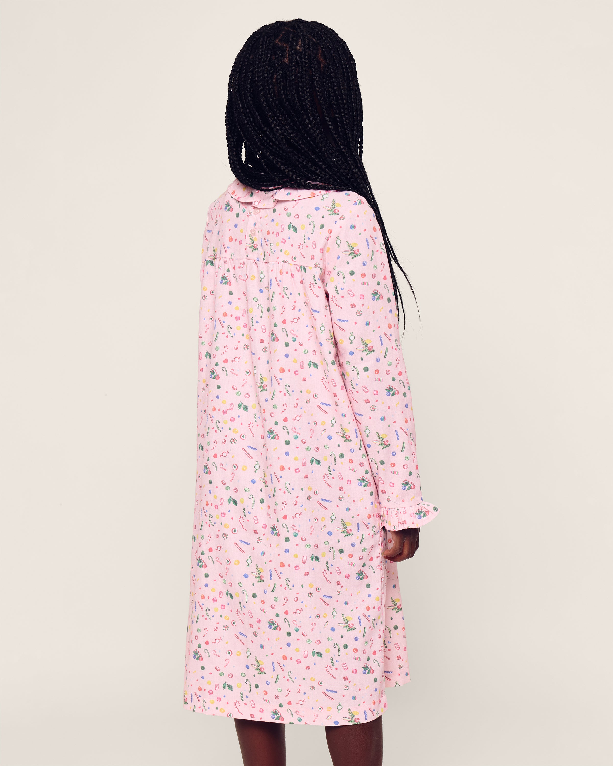 A person with long braided hair wears the Petite Plume Girls Twill Scarlett Nightgown in Vintage Sweets, a light pink, flame-retardant dress adorned with colorful floral and object patterns, against a plain cream background.