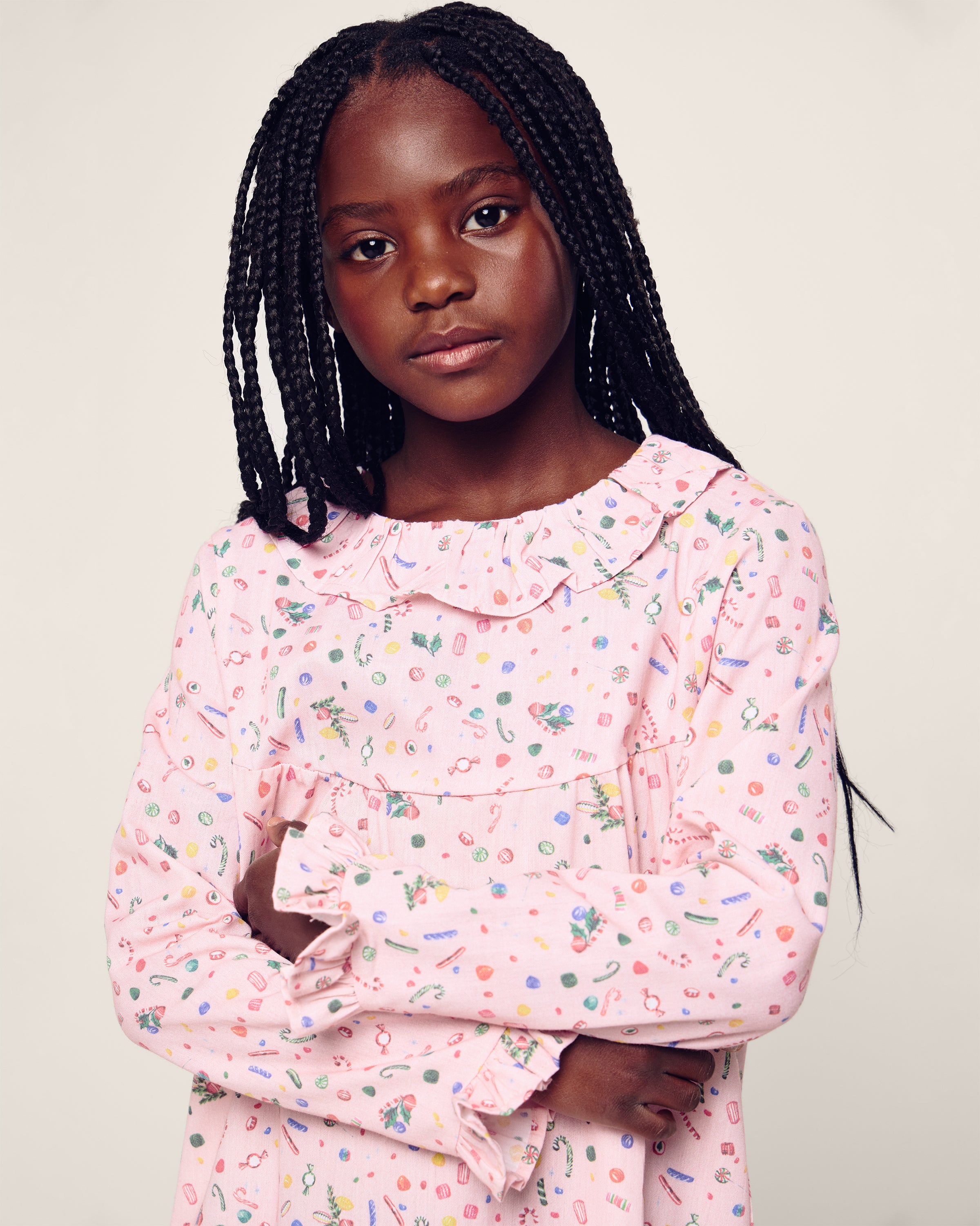 A young girl with long braided hair in a Petite Plume Girls Twill Scarlett Nightgown in Vintage Sweets stands against a neutral background, her arms gently crossed and expression calm and attentive.