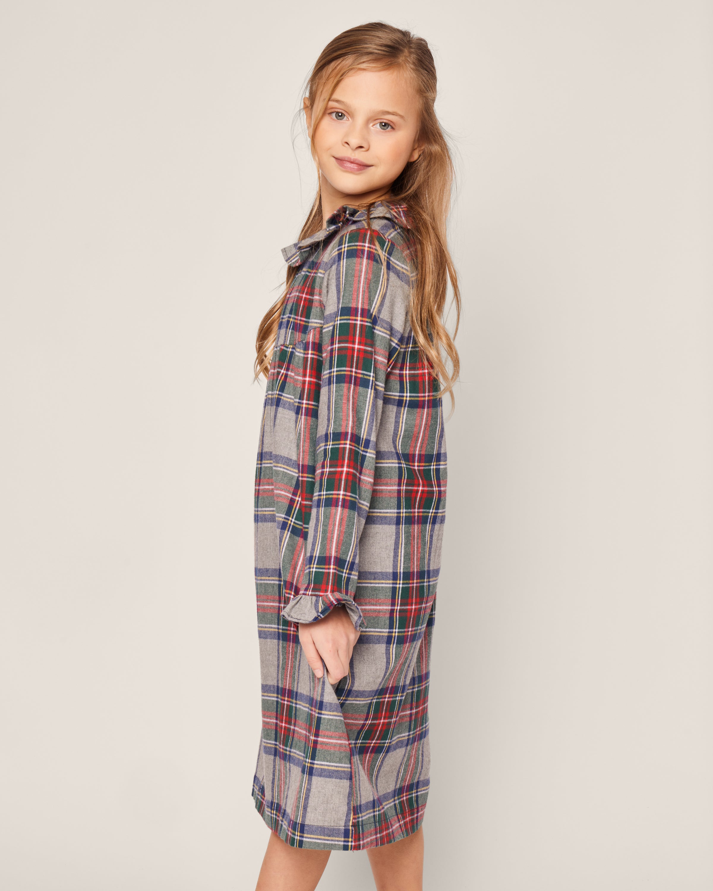 A young girl in Petite Plumes long-sleeved Girls Brushed Cotton Scarlett Nightgown in Westminster Tartan smiles slightly while standing sideways against a plain background.