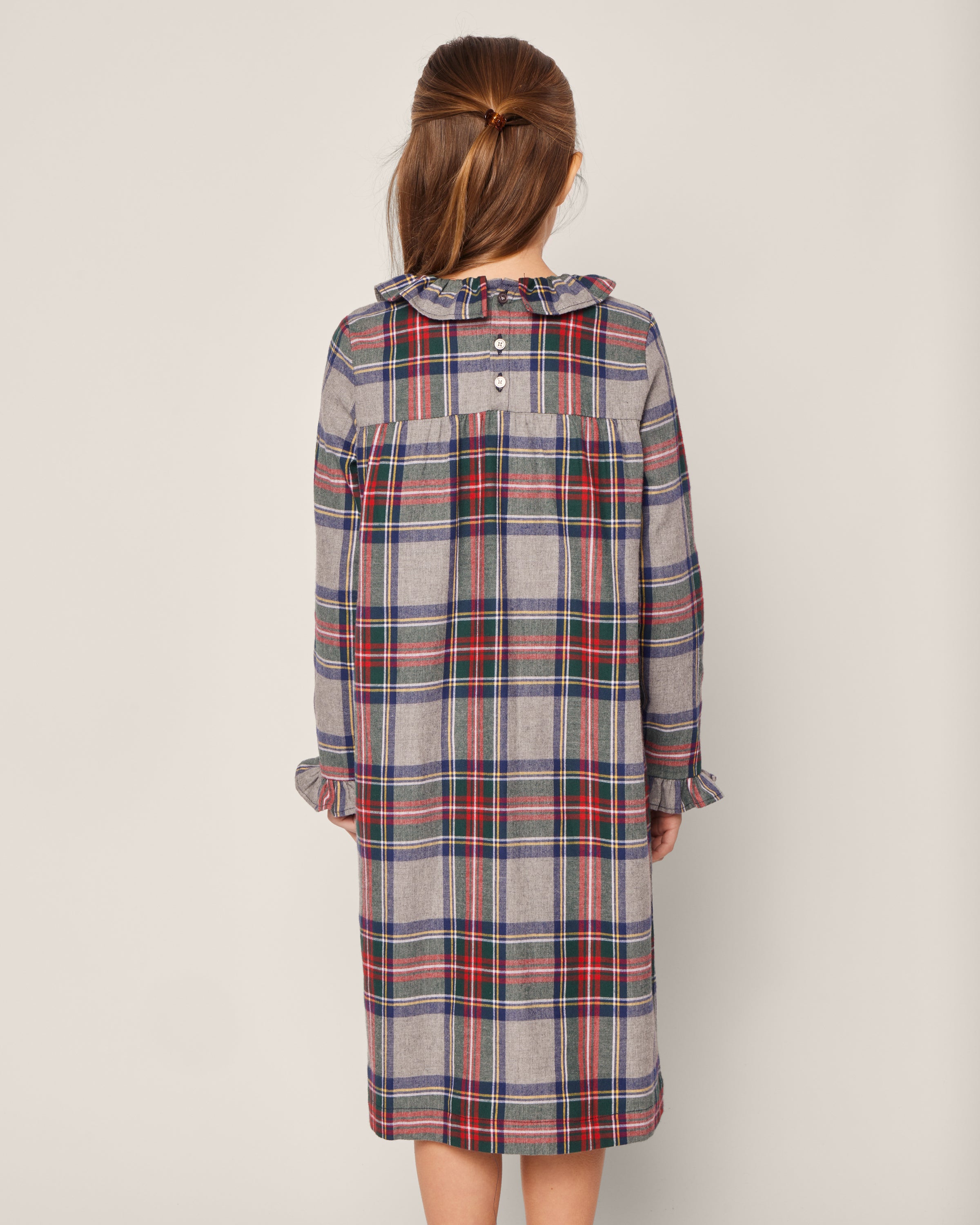 A young girl with long hair stands facing away from the camera, wearing Petite Plumes Girls Brushed Cotton Scarlett Nightgown in Westminster Tartan. The plaid dress has ruffles at the collar and cuffs, with a blue, red, green, and yellow design featuring two upper back buttons for a snug fit.