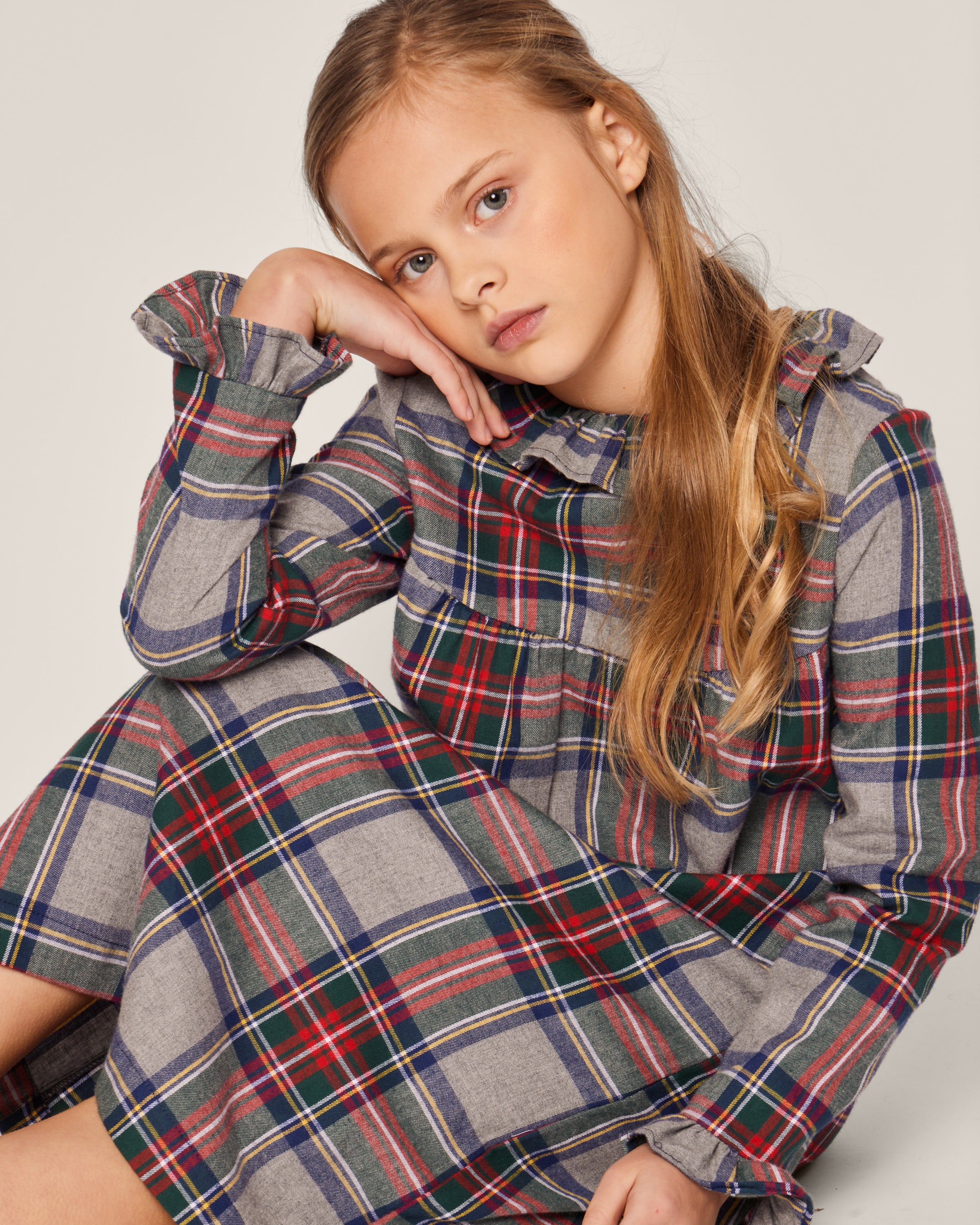 A young blonde girl thoughtfully rests her head on her hand, wearing a Petite Plume Girls Brushed Cotton Scarlett Nightgown in Westminster Tartan with gray, red, green, and blue hues, gazing at the camera against a plain light background.