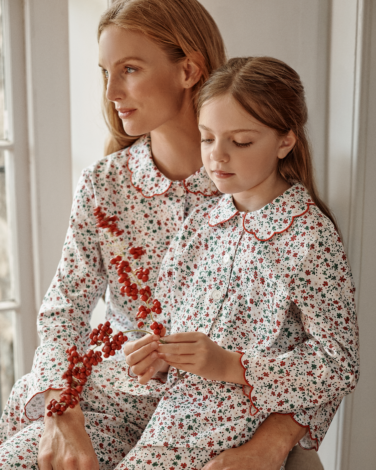 A mother with auburn hair and her young daughter sit together indoors, both dressed in matching Kid’s Twill Colette Pajama Sets in Winter Garden—a white, long-sleeved set featuring a red and green floral print with scalloped red trim. The girl holds a sprig of bright red berries.
