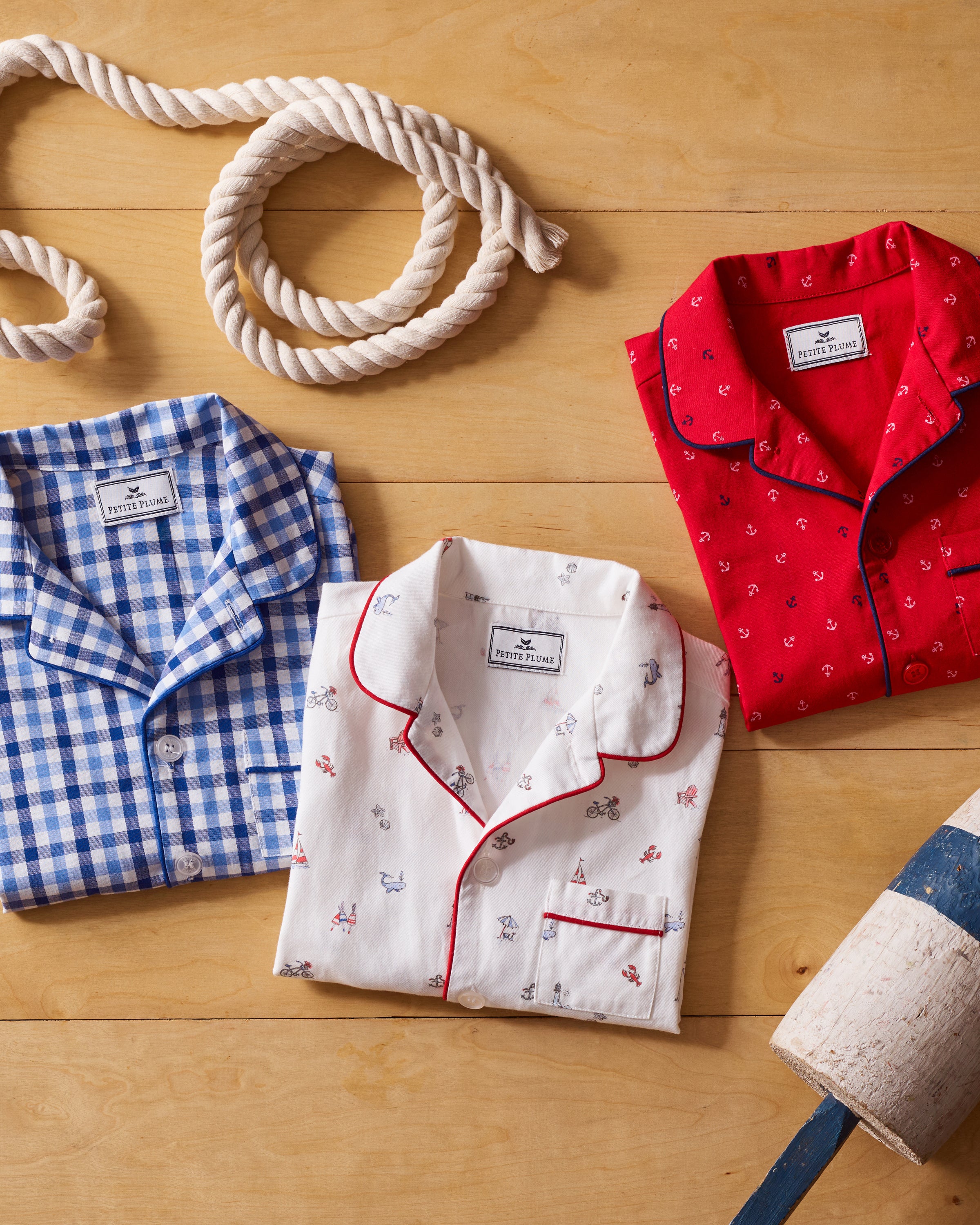 Three folded Kids Twill Pajamas from Petite Plume are displayed on a wooden surface: a blue checkered, a white with nautical prints, and one with red anchor patterns. A coiled rope and nautical buoy complete this charming Coastal Charms pajama set scene.