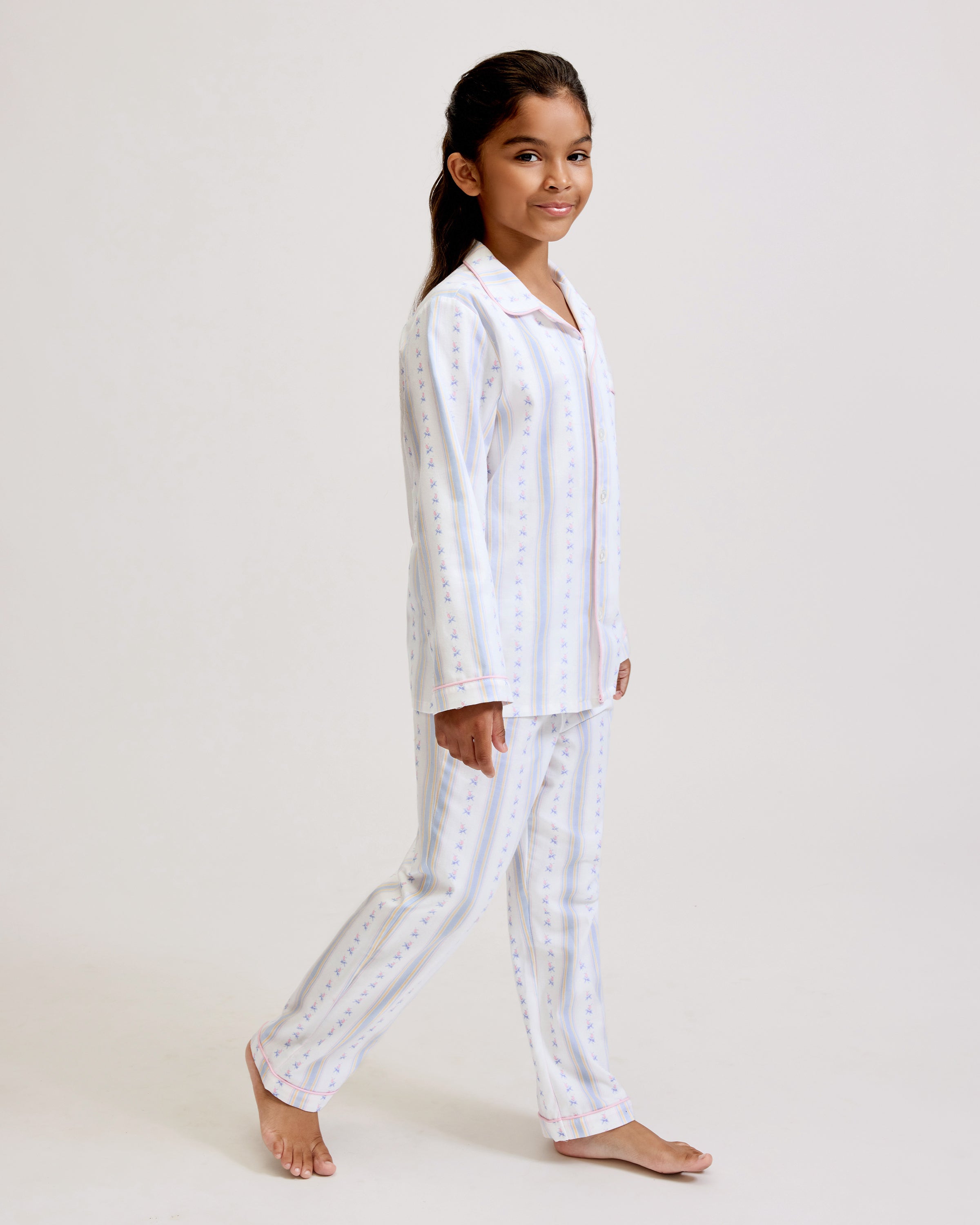 A young girl with dark hair stands barefoot on a plain background, wearing the Kid's Twill Pajama Set in Cottage Stripe—matching white pajamas with pastel stripes and a chest pocket—facing forward and smiling slightly.