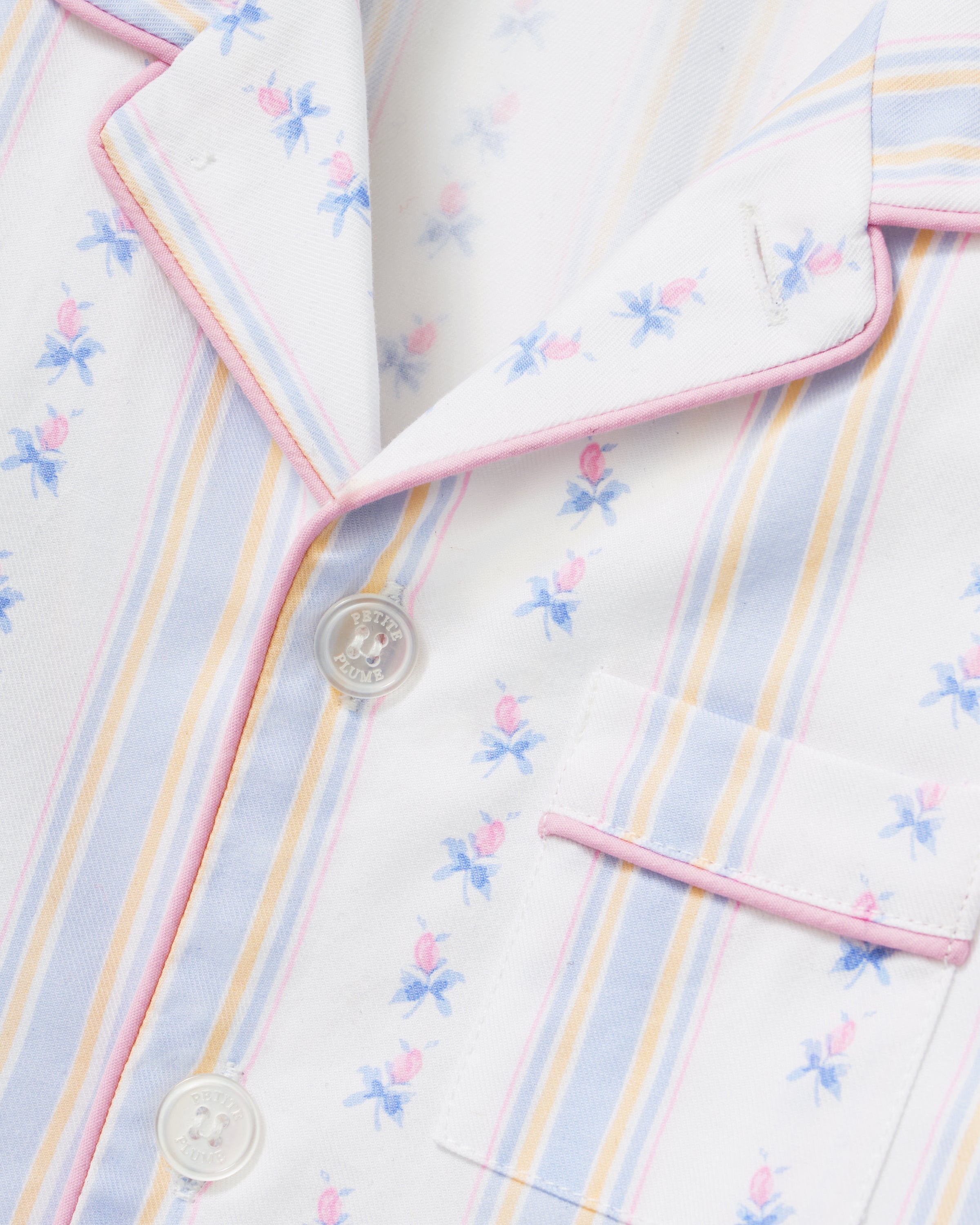 A young girl with dark hair stands barefoot on a plain background, wearing the Kid's Twill Pajama Set in Cottage Stripe—matching white pajamas with pastel stripes and a chest pocket—facing forward and smiling slightly.