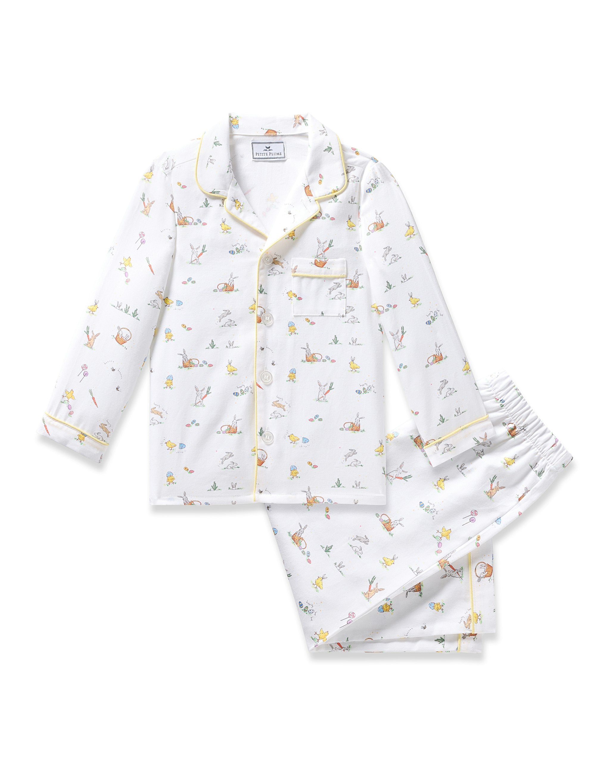 A young girl with long dark hair in a braid smiles barefoot, wearing the Kid's Twill Pajama Set in Easter Frolic, against a plain light background.