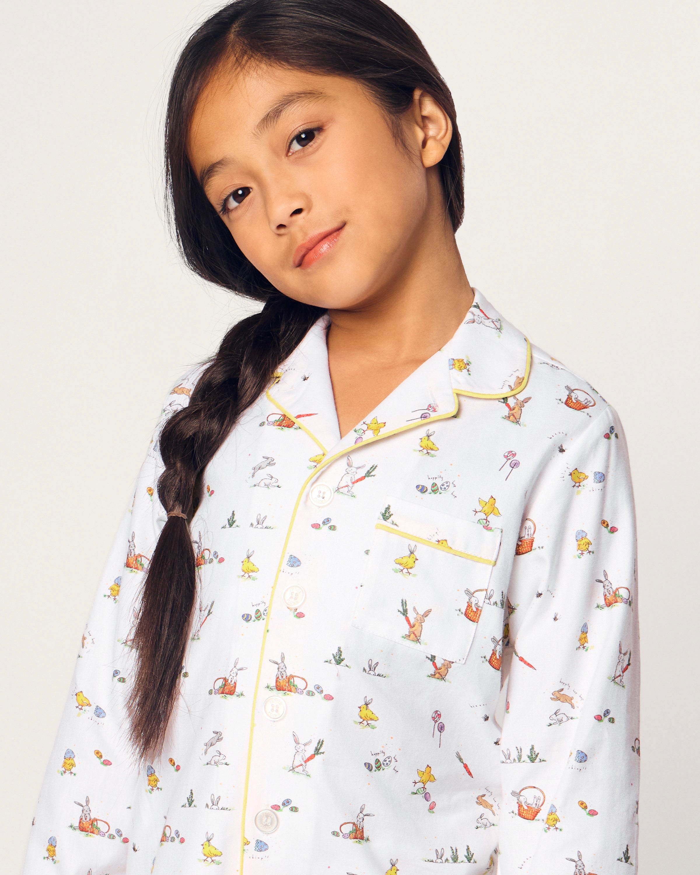A young girl with long dark hair in a braid smiles barefoot, wearing the Kid's Twill Pajama Set in Easter Frolic, against a plain light background.