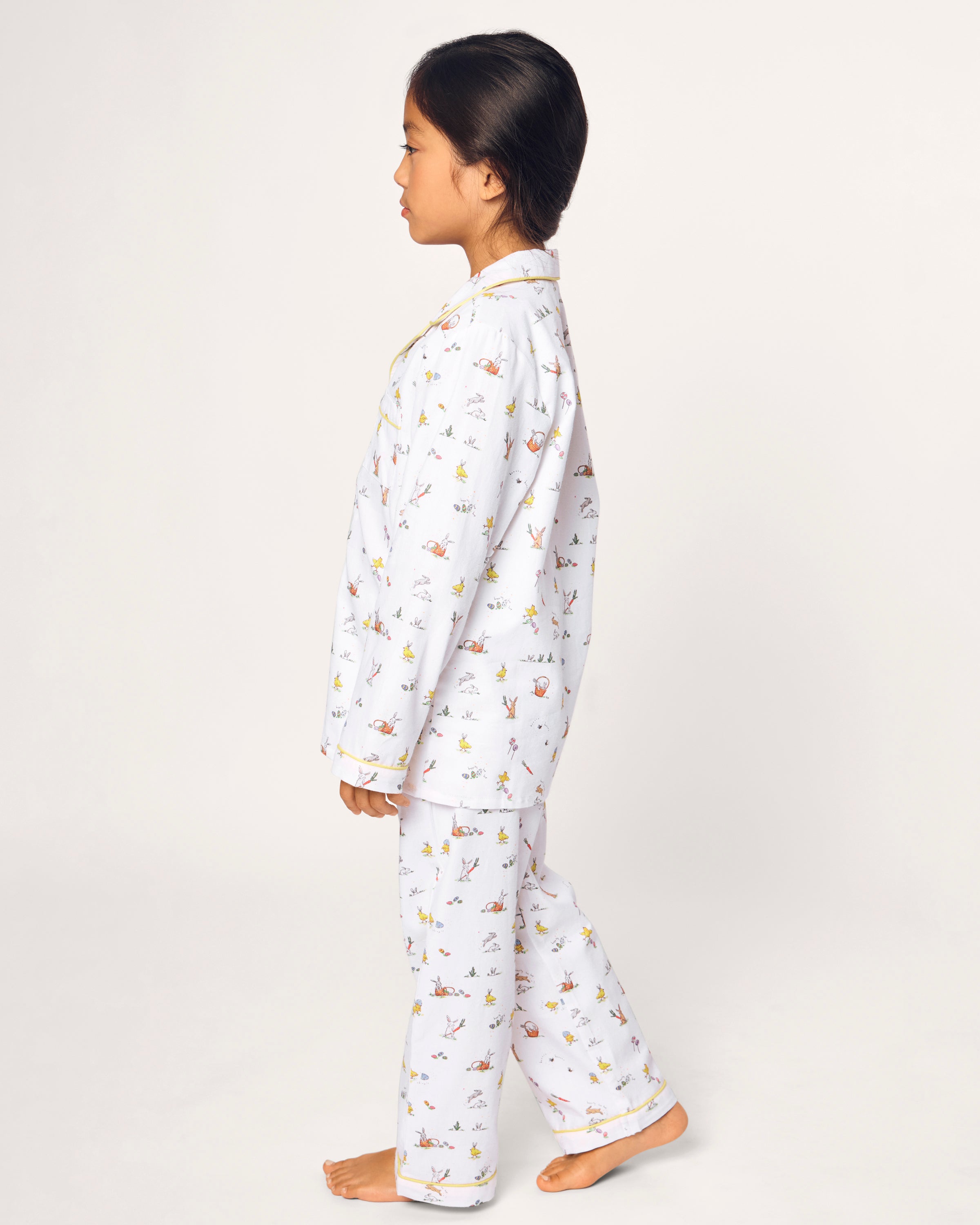 A young girl with long dark hair in a braid smiles barefoot, wearing the Kid's Twill Pajama Set in Easter Frolic, against a plain light background.