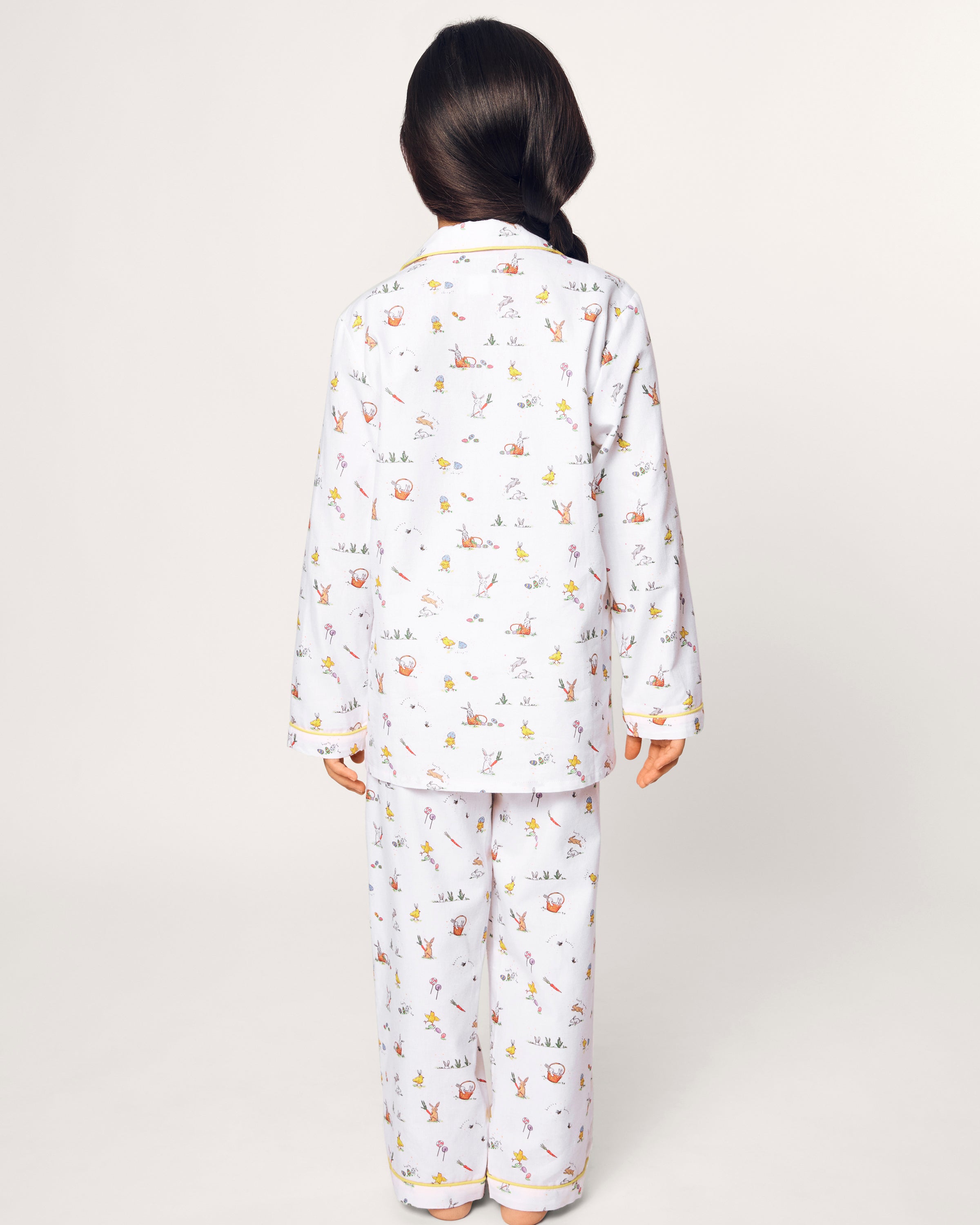 A young girl with long dark hair in a braid smiles barefoot, wearing the Kid's Twill Pajama Set in Easter Frolic, against a plain light background.