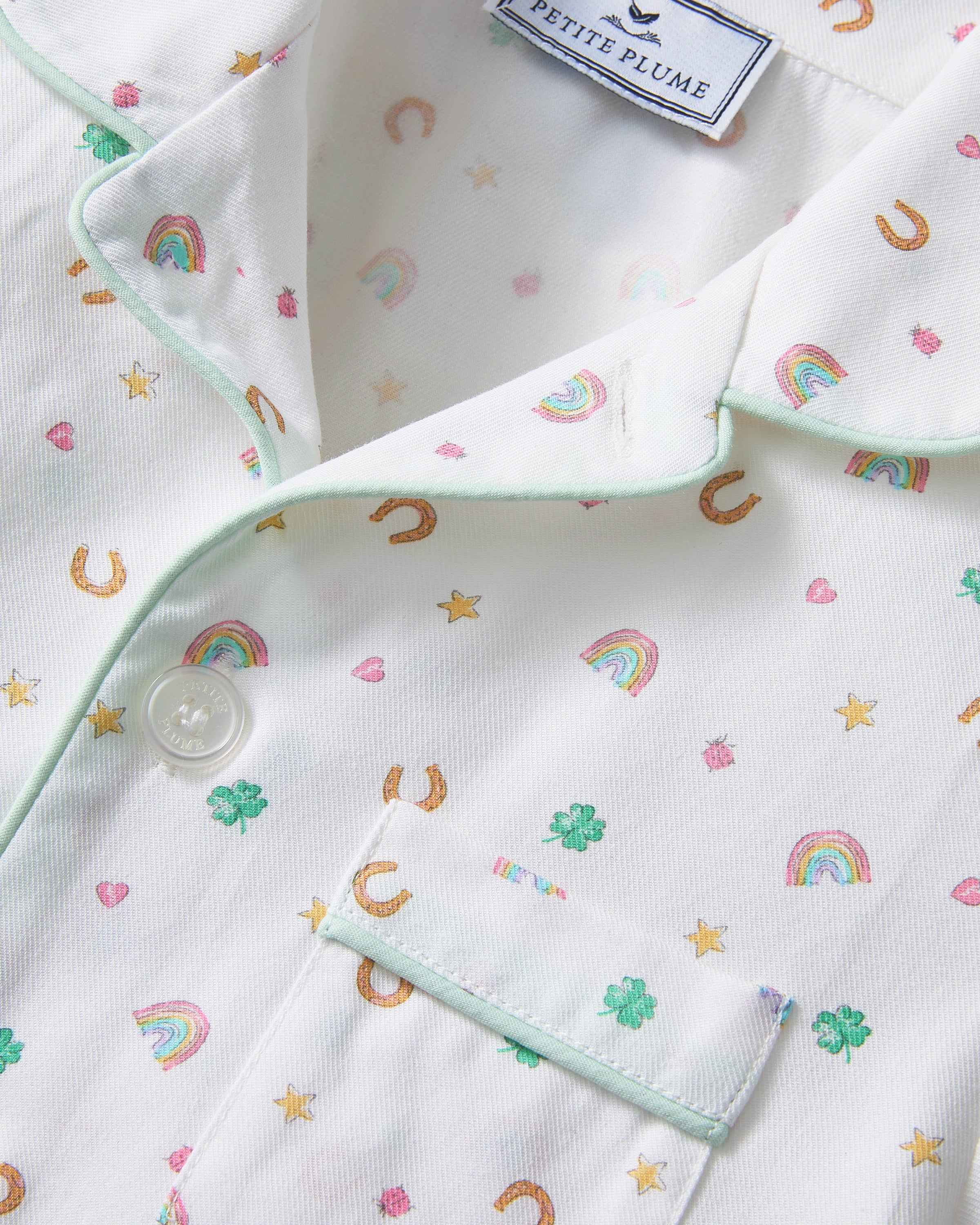 A child with short curly hair stands facing forward, wearing the Kid's Twill Pajama Set in Good Luck Charms—a white button-up pajama with a colorful pattern and matching pants—against a plain light background.
