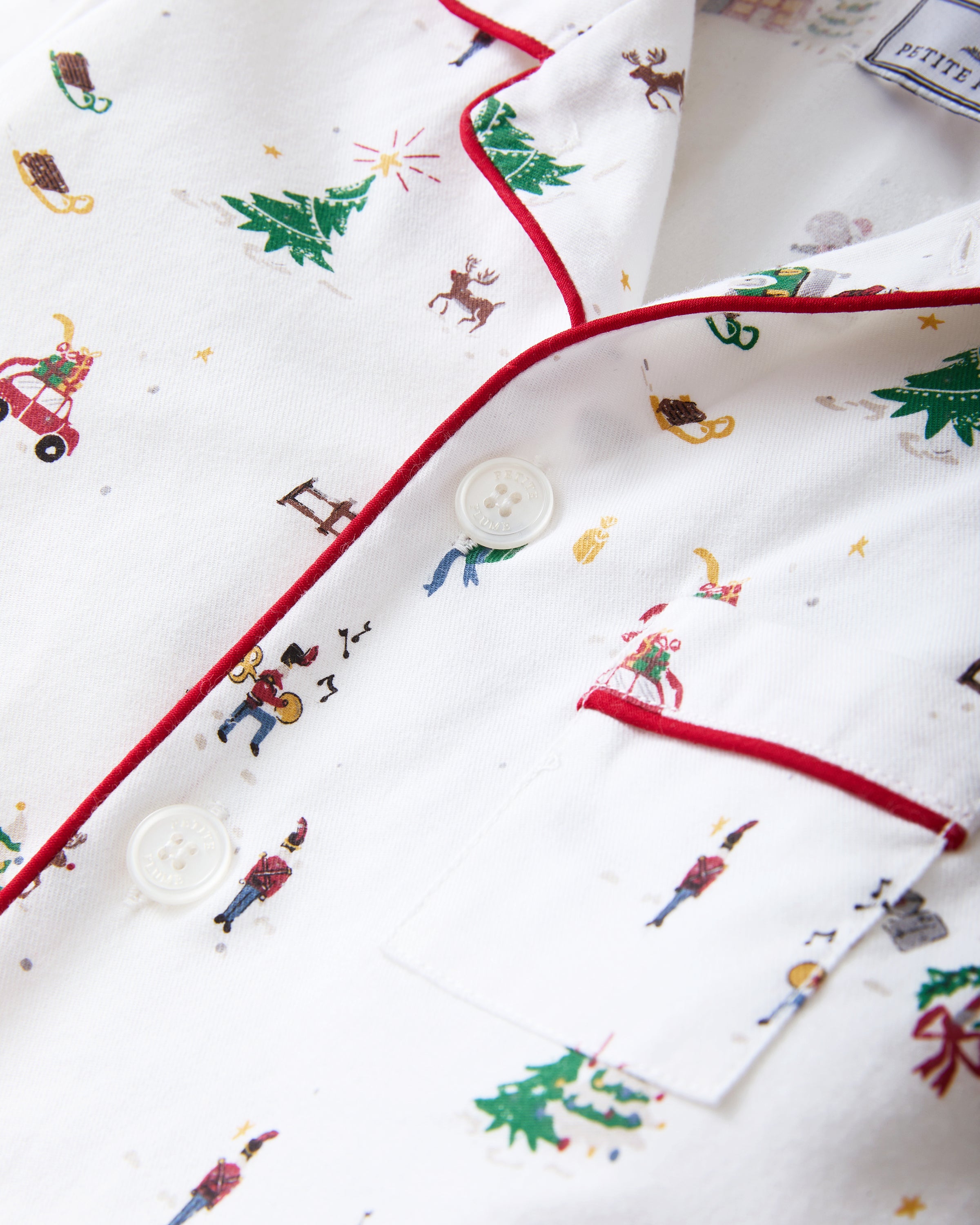 The Kid's Twill Pajama Set in Holiday Town features white long-sleeve, button-up pajamas with red piping and a festive town print of trees, dogs, people, cars, and houses; pants and top are neatly arranged.
