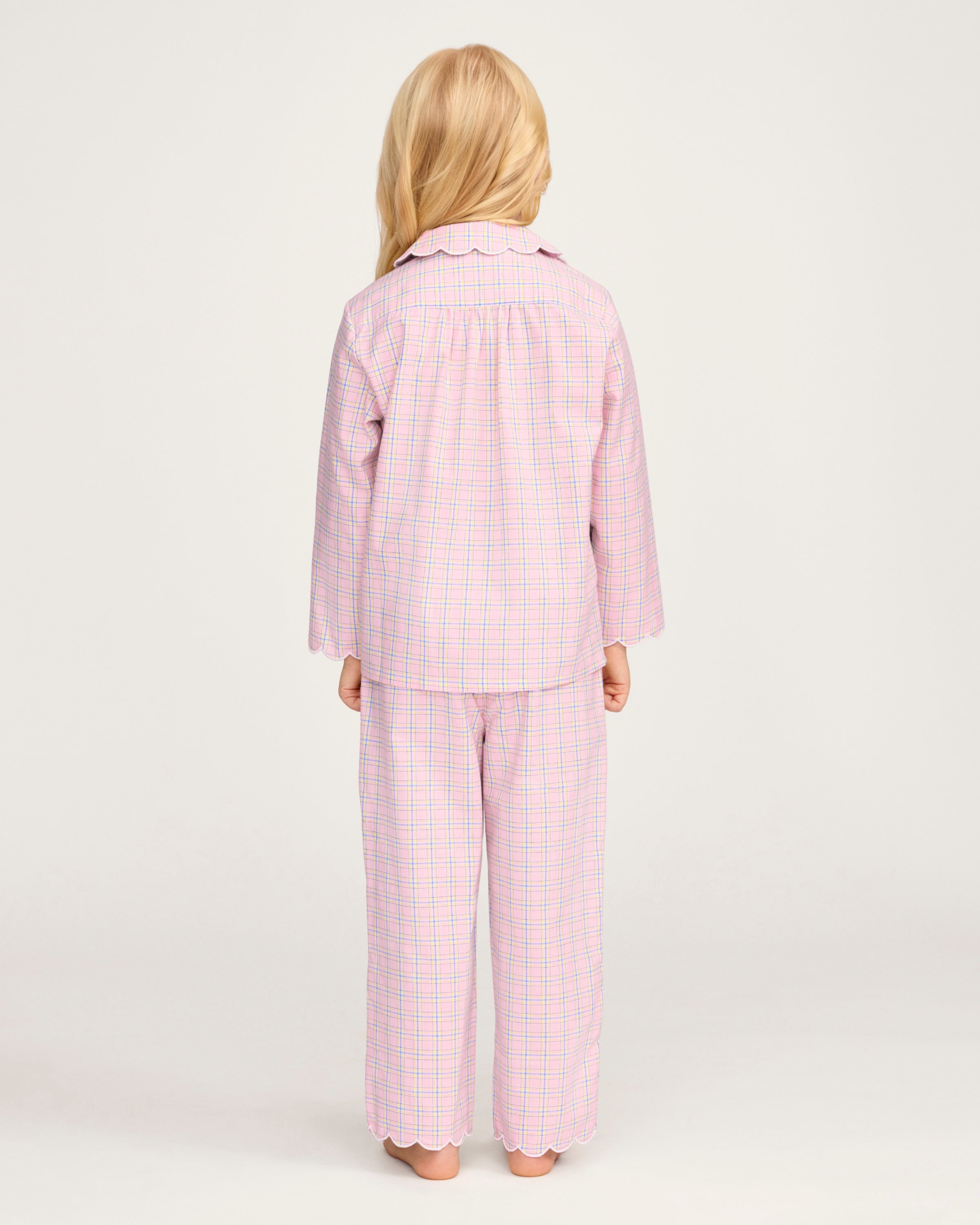 Young girl with long blonde hair wears the Kid's Twill Colette Pajama Set in Peony Plaid, featuring pink and white checks, scalloped edges, and stands barefoot against a plain light background.