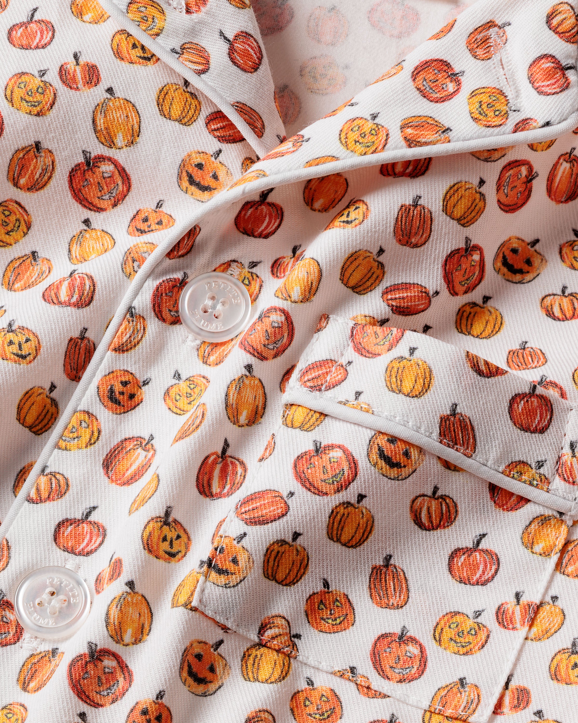 Close-up of Petite Plumes Kids Twill Pajama Set in Pumpkin Patch, featuring a white fabric with orange pumpkins and jack-o-lanterns. The design includes visible buttons and a pocket, indicating flame-retardant sleepwear suitable for kids.