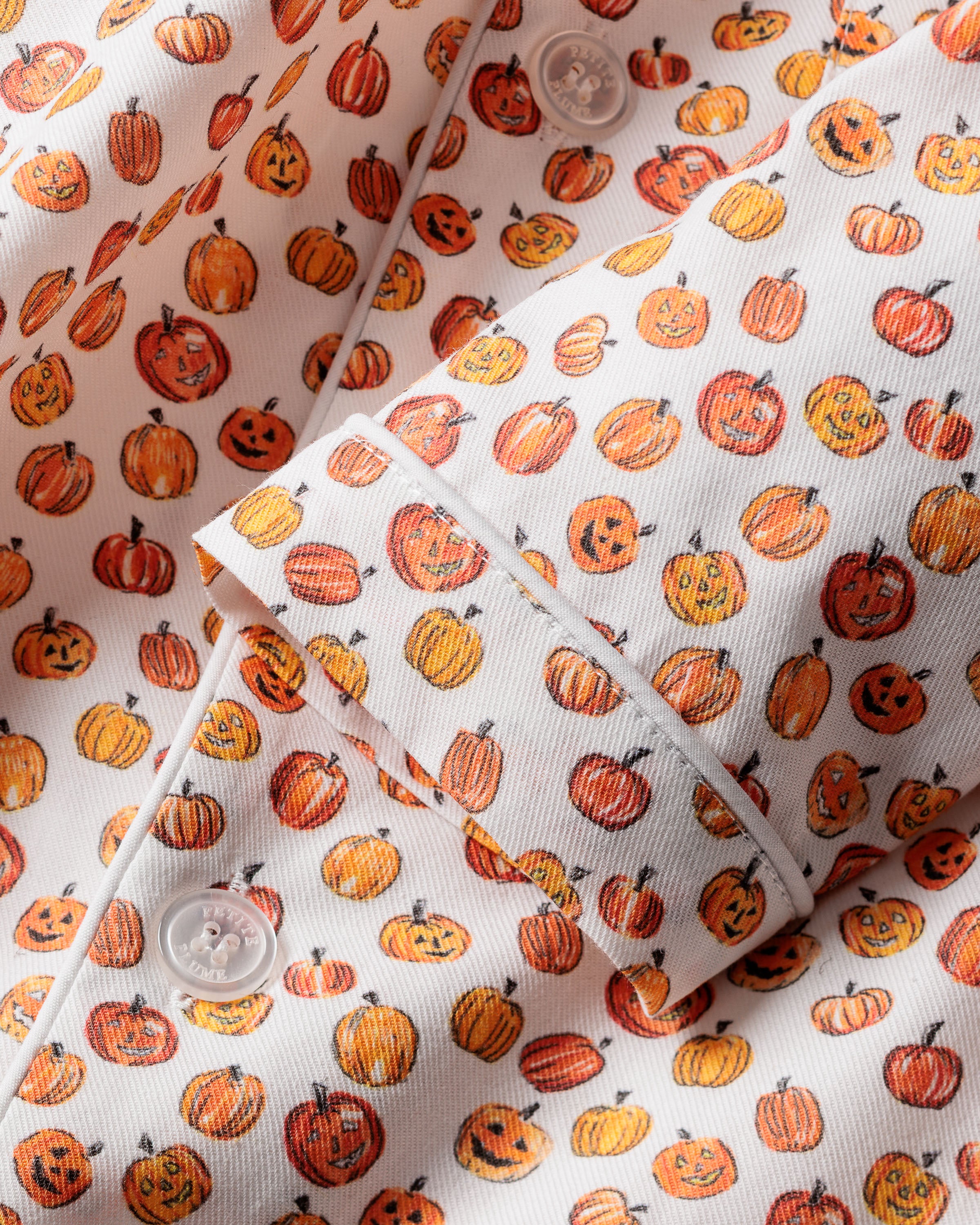 The Kids Twill Pajama Set in Pumpkin Patch by Petite Plume features a playful kids’ print with small, smiling pumpkins in orange and yellow. It includes white buttons and a textured weave, creating a lively Halloween-themed design perfect for flame-retardant sleepwear.
