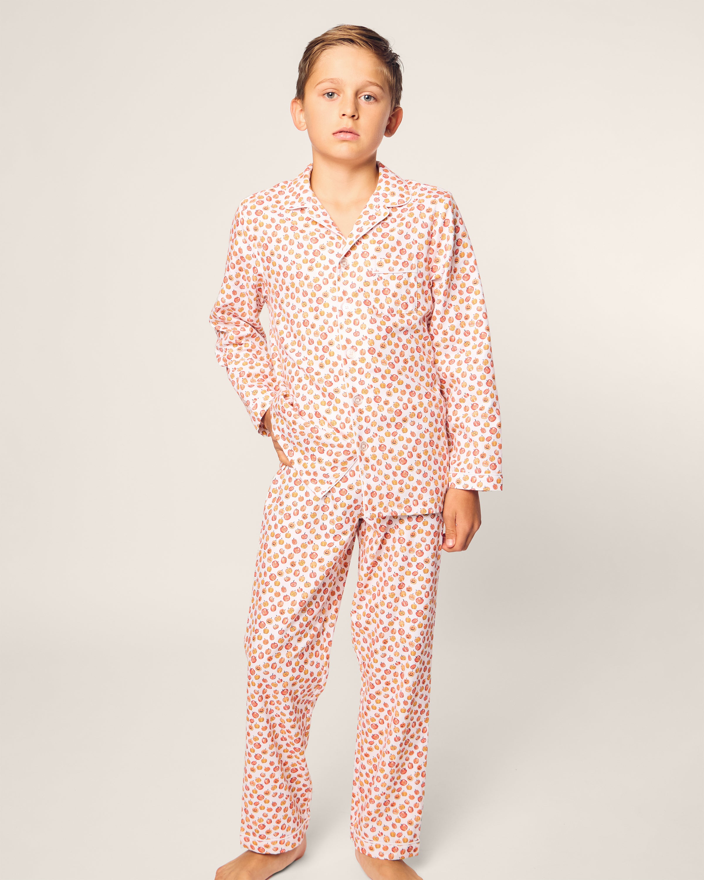 A child barefoot against a plain background, wearing Petite Plumes Kids Twill Pajama Set in Pumpkin Patch, light-colored and flame-retardant with small red and yellow kids pattern.