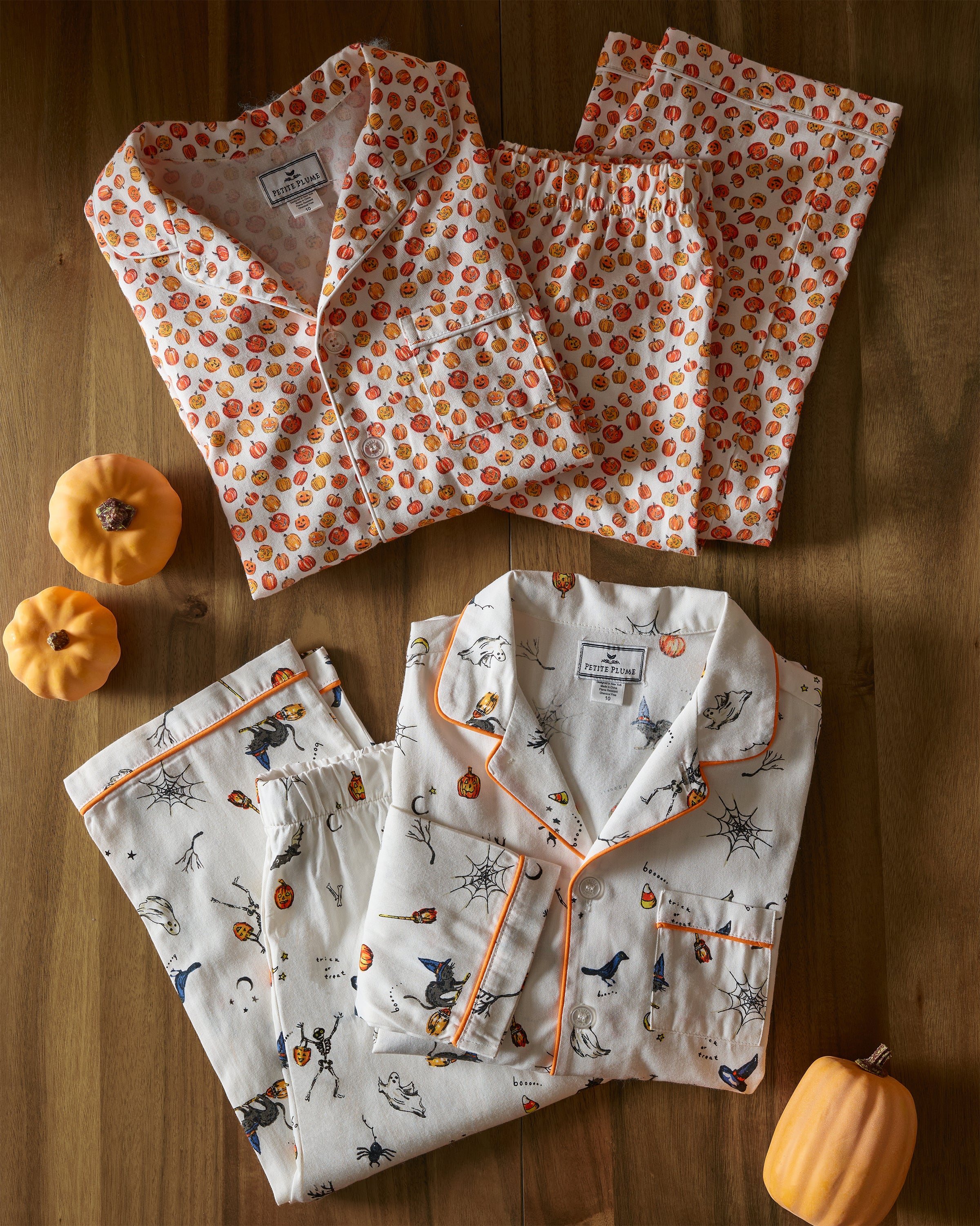 Two Petite Plume Kids Twill Pajama Sets in Pumpkin Patch are displayed on wood. One features pumpkins, the other has witches and spiders. Three small pumpkins are artfully placed around these cozy, chemical-free sleepwear sets for a safe Halloween night.