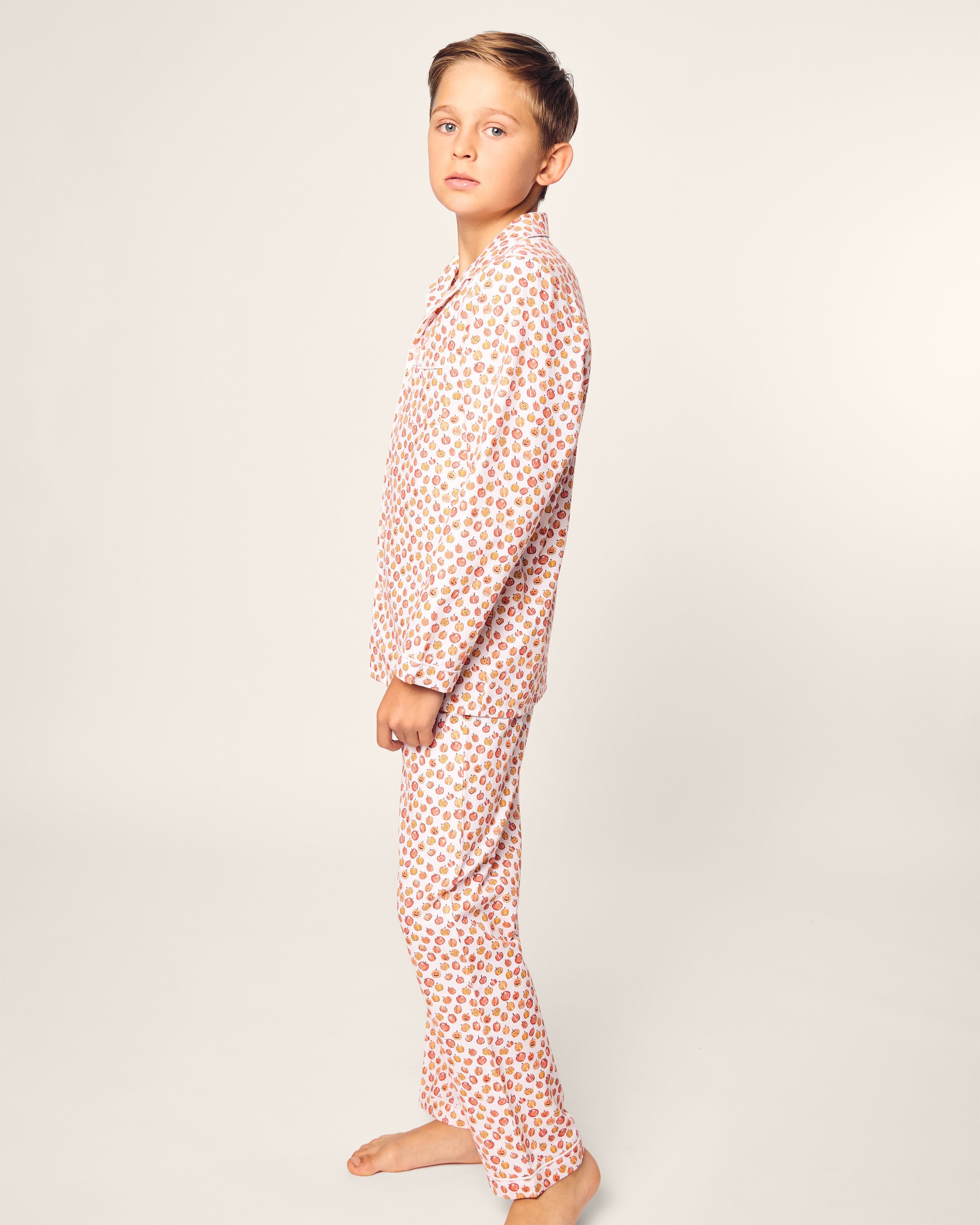 A young boy in Petite Plumes Kids Twill Pajama Set in Pumpkin Patch—featuring a red and gold geometric pattern—stands barefoot and sideways against a plain background. The flame-retardant sleepwear ensures safety as he looks at the camera with a neutral expression.