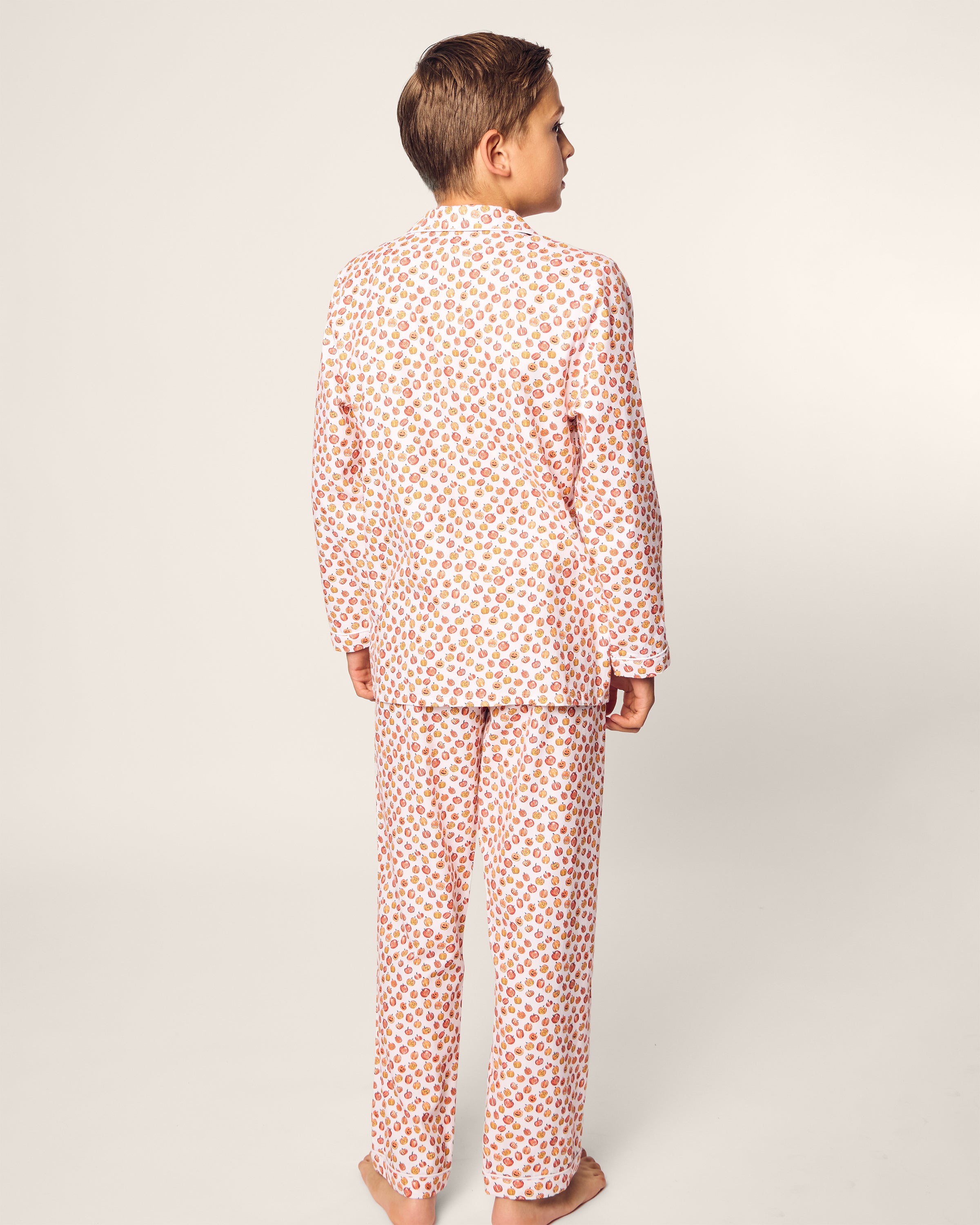 A young boy stands facing away, dressed in Petite Plumes Kids Twill Pajama Set in Pumpkin Patch. This flame-retardant sleepwear includes a long-sleeved top and pants. The neutral background highlights the playful yet safe design of his cozy ensemble.