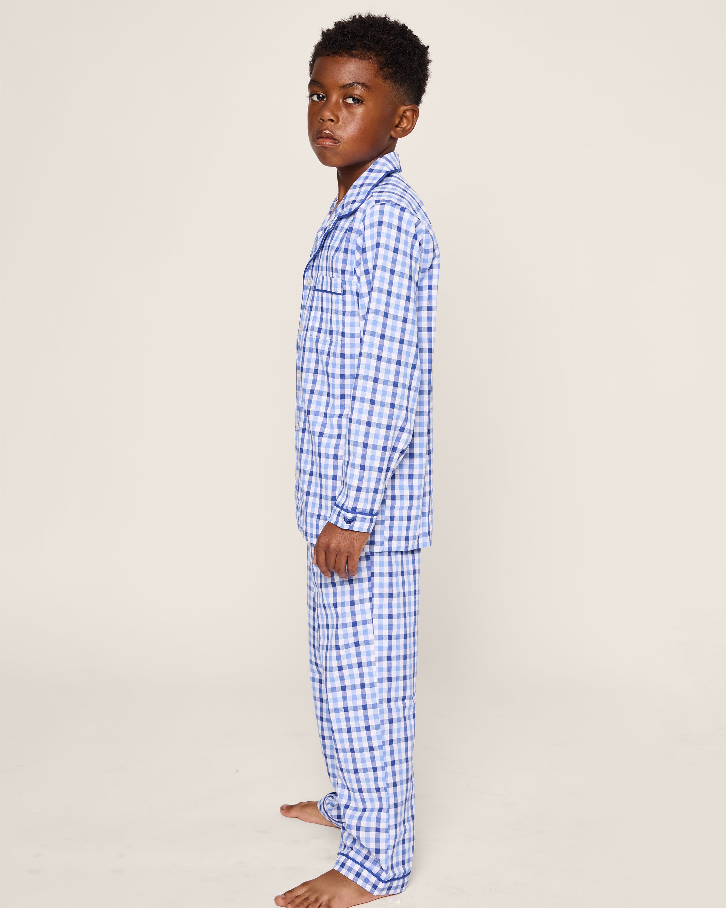 A young boy stands barefoot, sideways in Petite Plumes Kids Twill Pajama Set in Royal Blue Gingham. He looks neutrally at the camera against a plain off-white background.