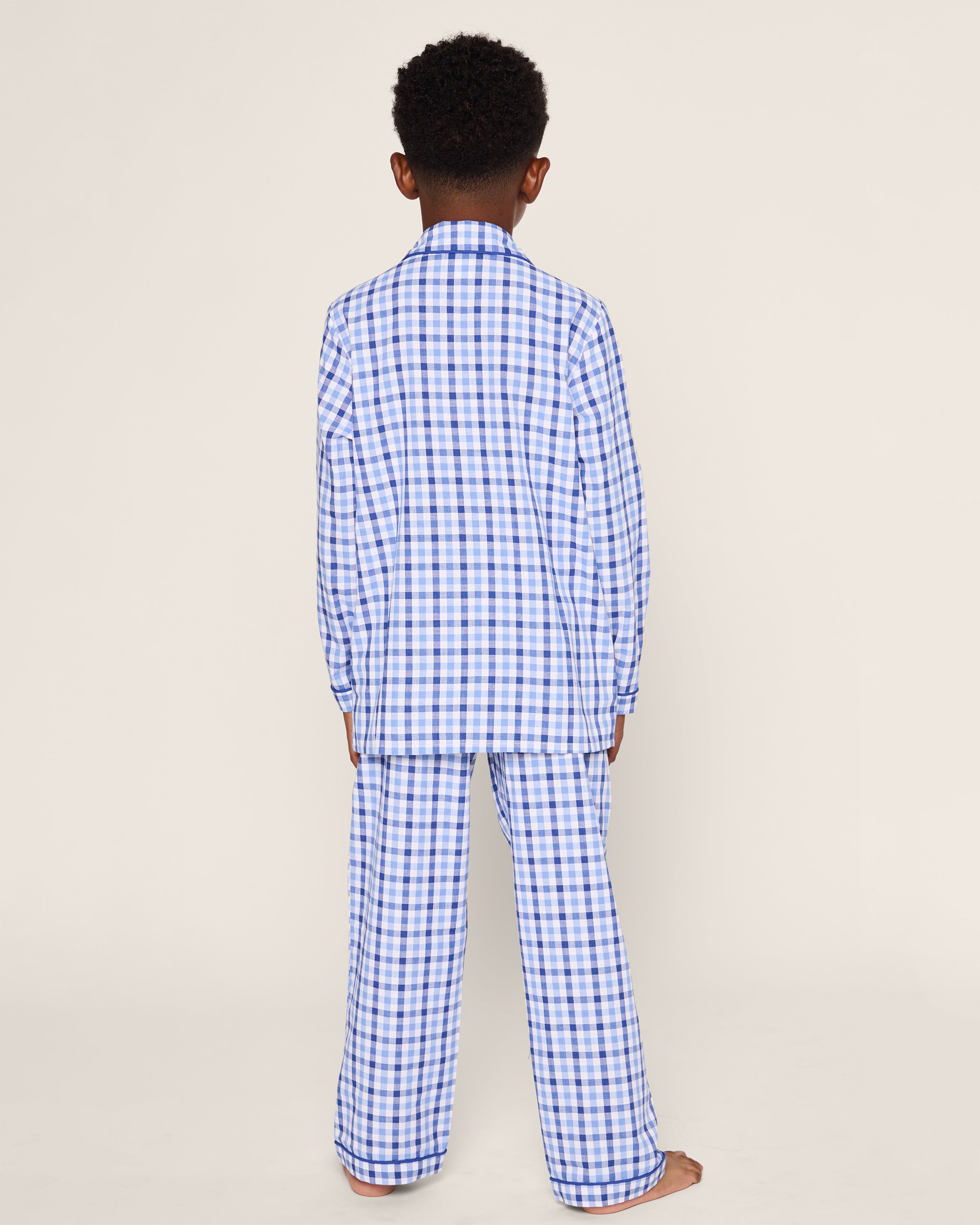 A child stands with their back turned, wearing Petite Plumes Kids Twill Pajama Set in Royal Blue Gingham. The background is a plain light gray.