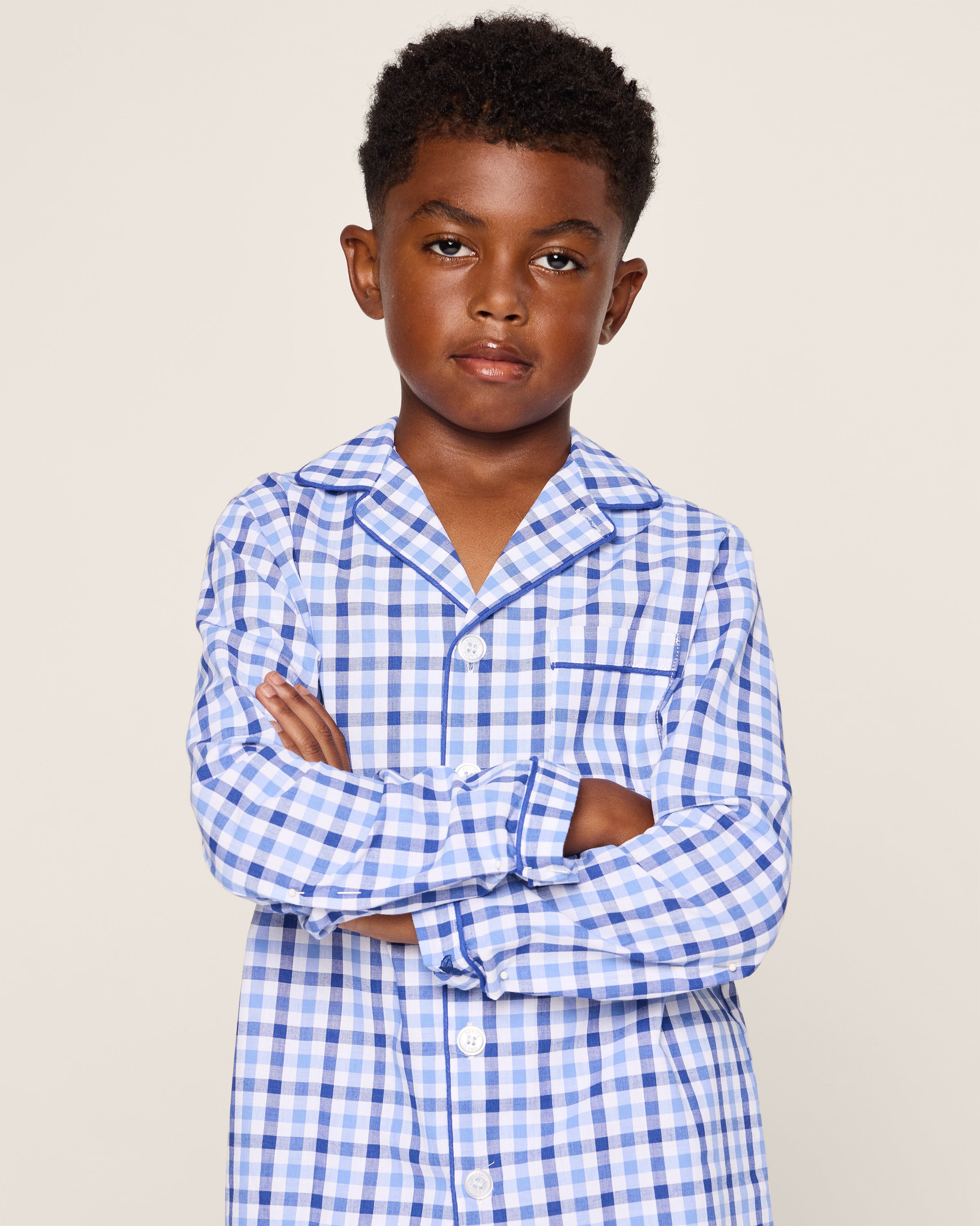 A young boy with short curly hair wears Petite Plumes Kids Twill Pajama Set in Royal Blue Gingham. He stands with crossed arms against a plain background, looking directly at the camera with a neutral expression.