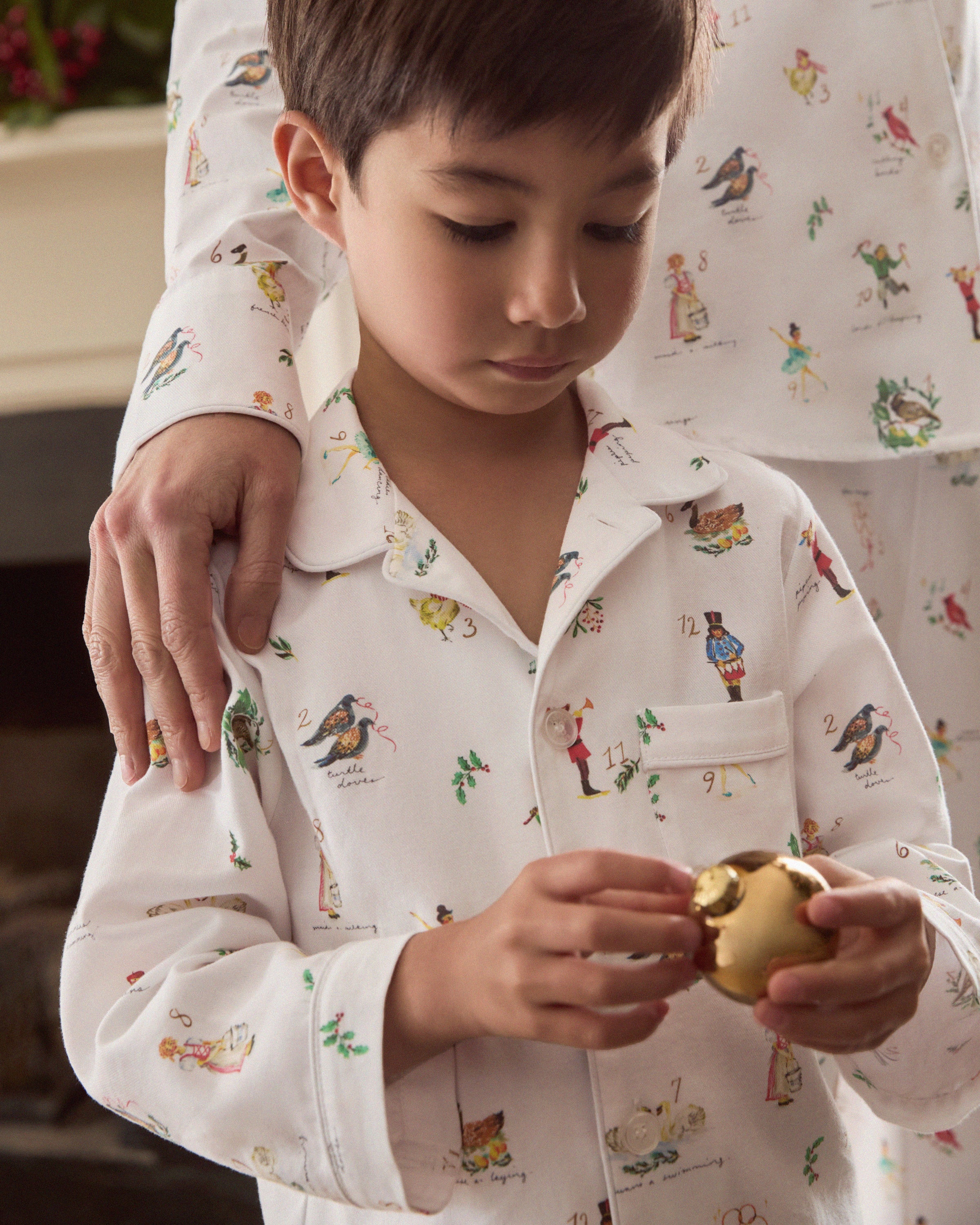 The Kid's Twill Pajama Set in 12 Days of Christmas features a white, long-sleeve button-up top with collar and front pocket, plus matching pants, all adorned with playful, colorful holiday illustrations of people and animals.