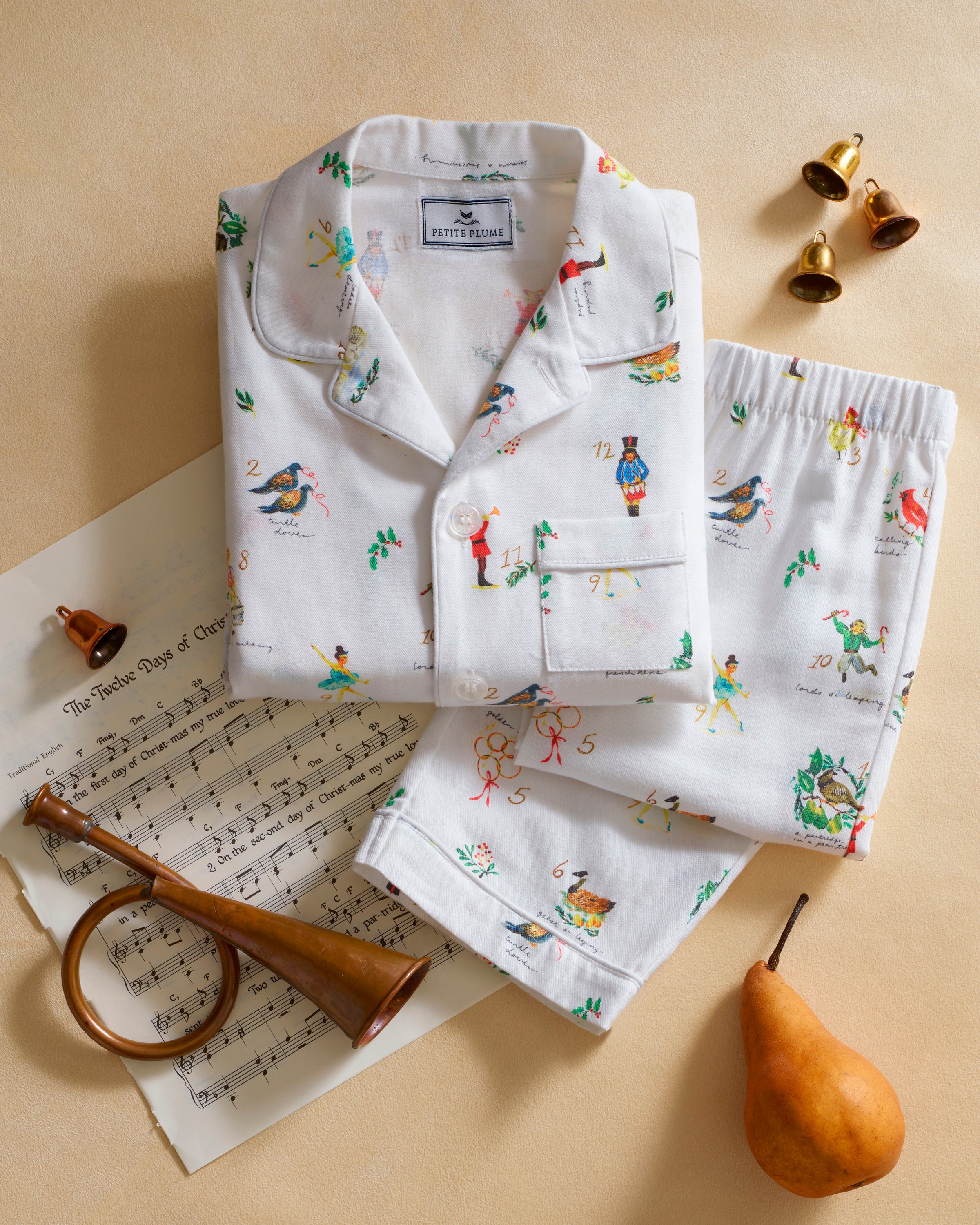 The Kid's Twill Pajama Set in 12 Days of Christmas features a white, long-sleeve button-up top with collar and front pocket, plus matching pants, all adorned with playful, colorful holiday illustrations of people and animals.