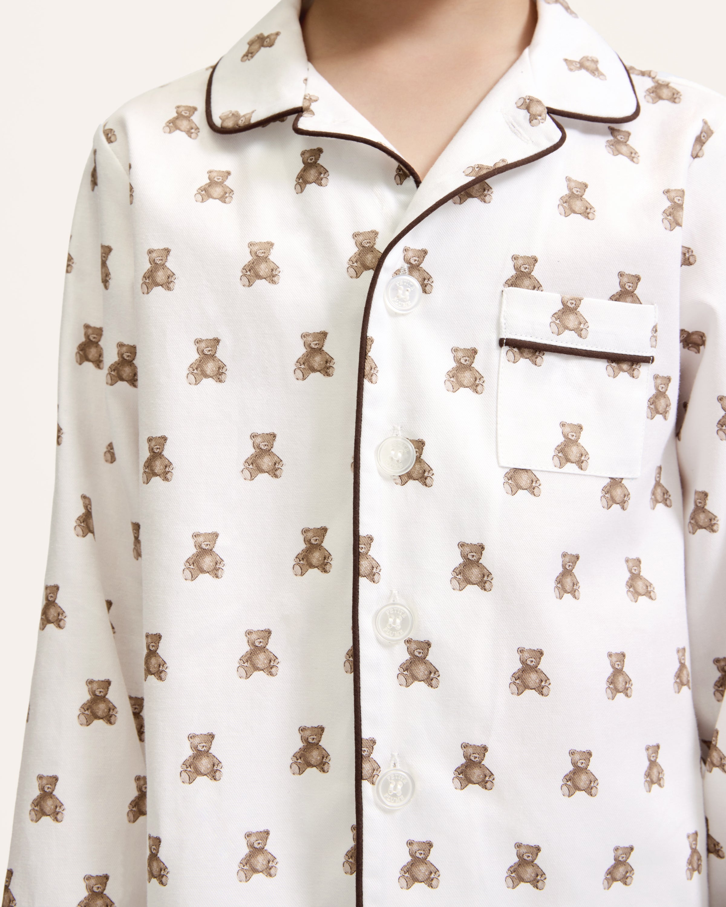 Kid's Twill Pajama Set in Teddy Tales: white long-sleeve, button-up pajamas with pants, featuring brown teddy bear print, dark piping along edges and chest pocket.