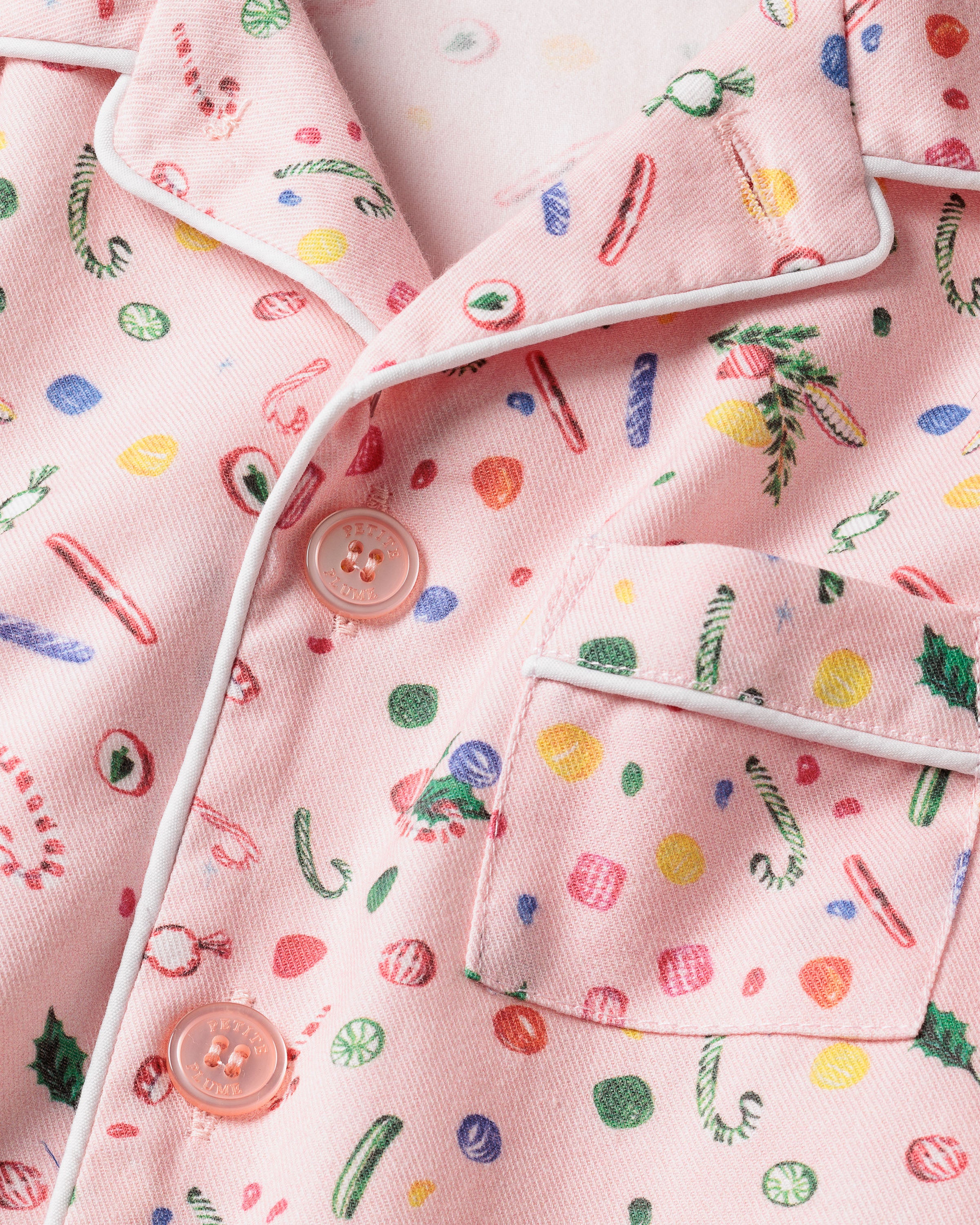 Close-up of Petite Plumes Kids Twill Pajama Set in Vintage Sweets, showcasing colorful holiday prints like candy canes, holly leaves, and ornaments. Features include a chest pocket, pink buttons, and white piping along the edges.