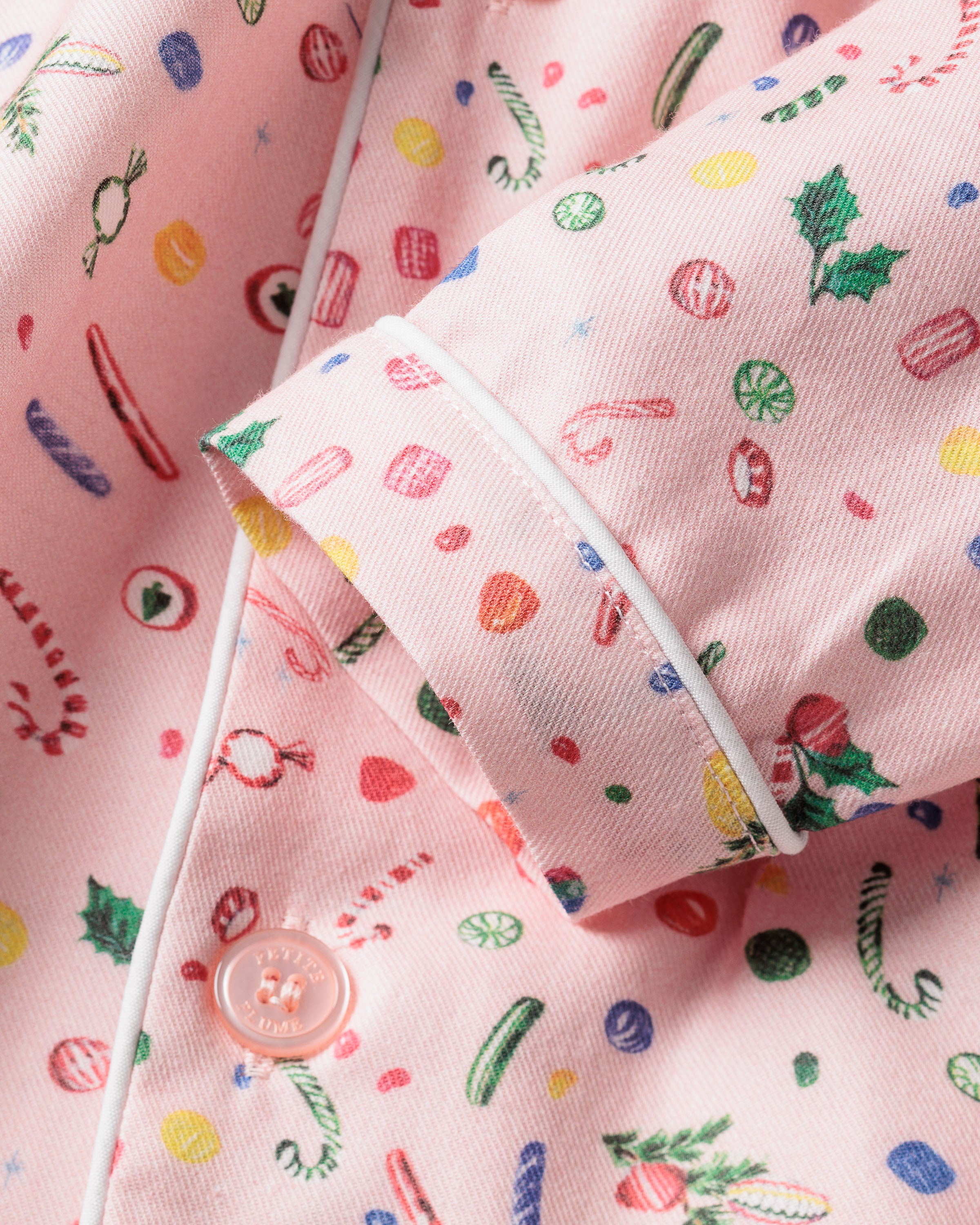 Close-up of Petite Plumes Kids Twill Pajama Set in Vintage Sweets, showing pink flame-retardant fabric with festive candy canes, holly leaves, and candy patterns. Details like a sleeve, button, and white piping highlight the twill design.