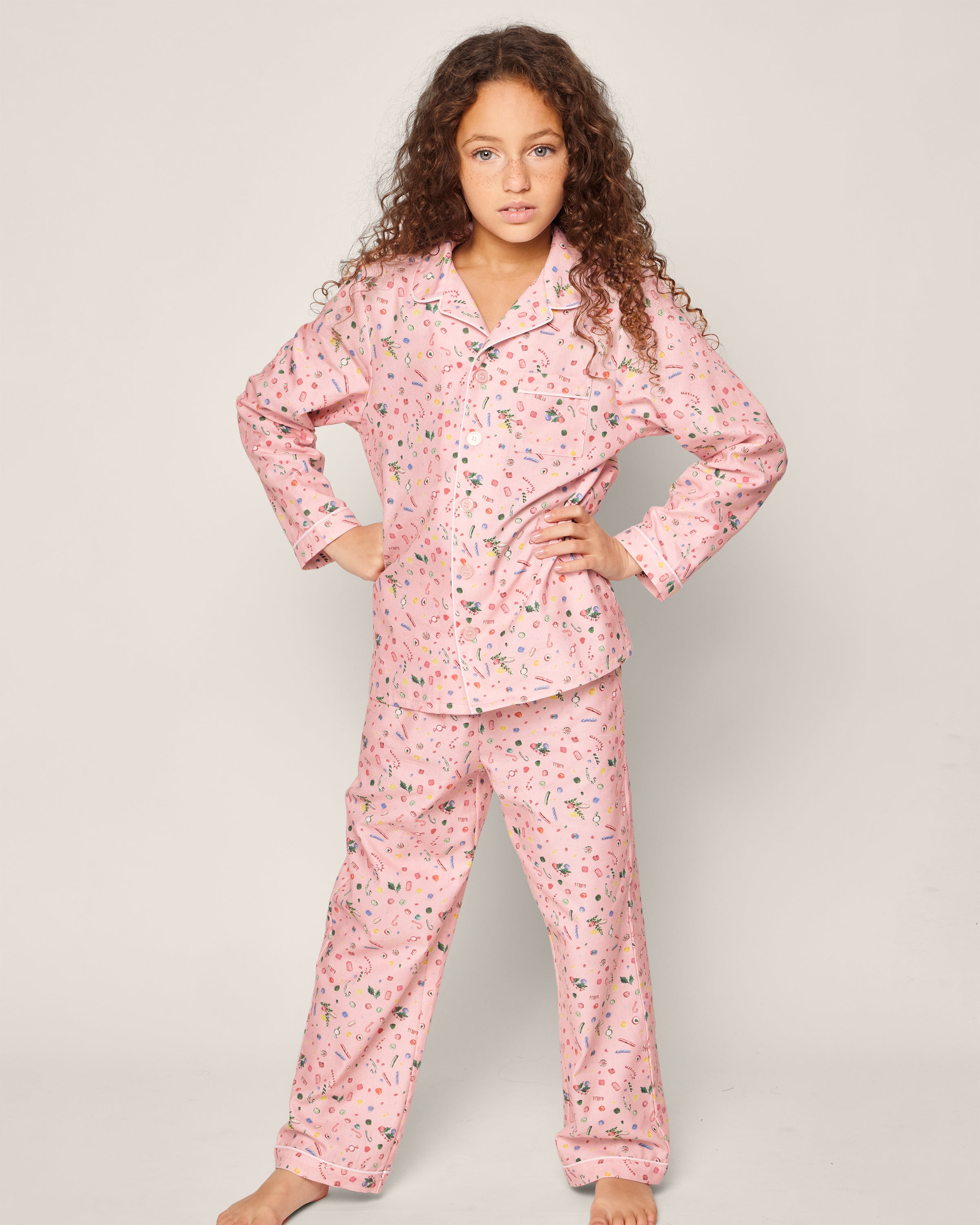 A young girl with long curly hair stands confidently in her Petite Plume Kids Twill Pajama Set in Vintage Sweets. She has her hands on her hips and is barefoot against a plain background.