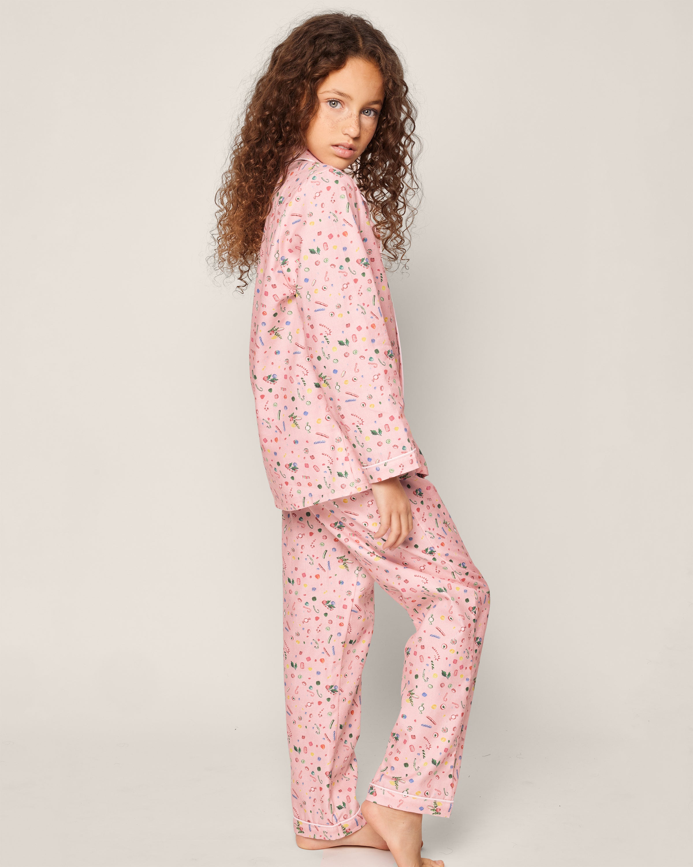 A child with curly brown hair stands barefoot in a Petite Plume Kids Twill Pajama Set in Vintage Sweets, adorned with small colorful patterns, looking back over their shoulder against a light background.
