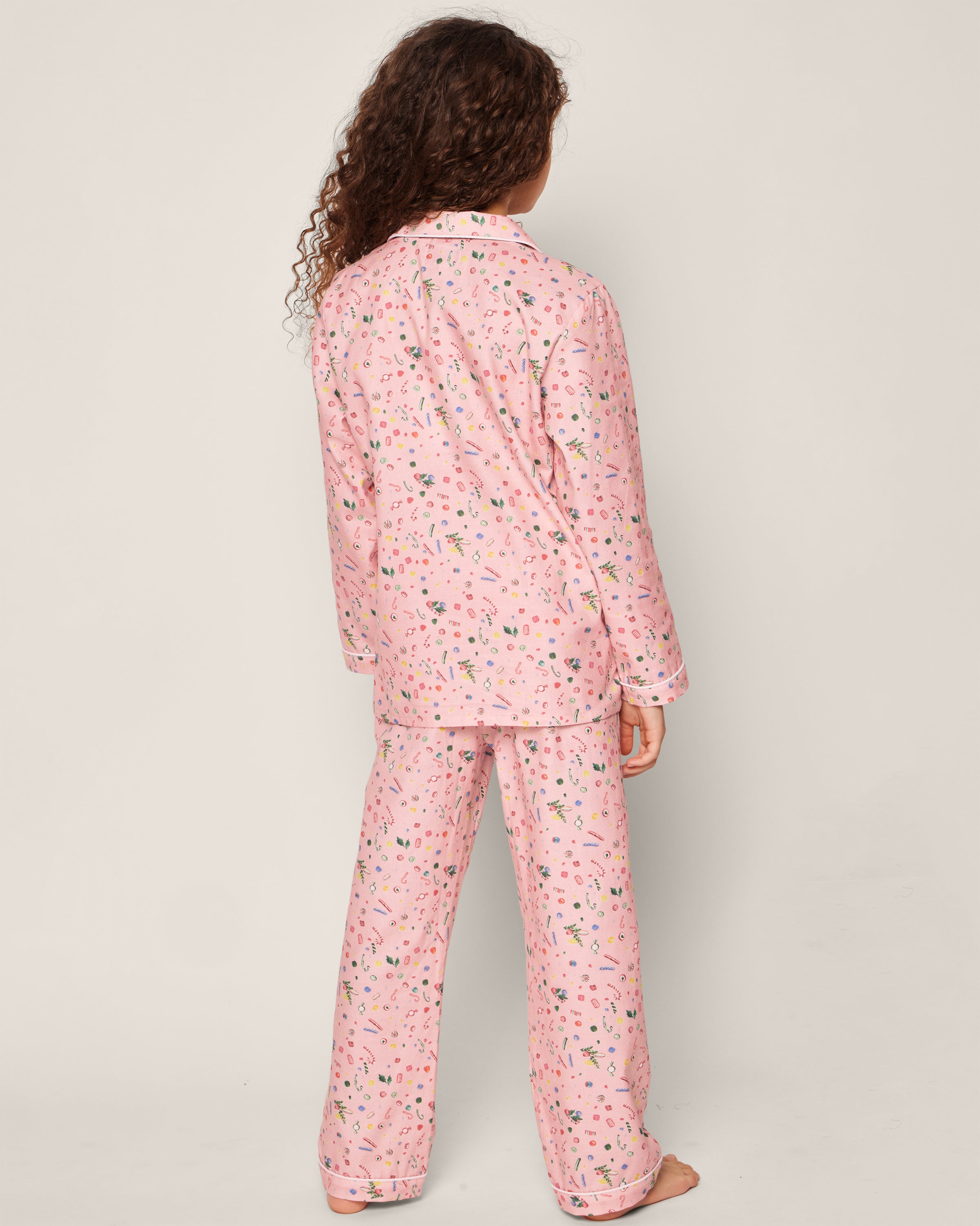 A child with curly hair stands facing away, wearing Petite Plumes Kids Twill Pajama Set in Vintage Sweets. The colorful patterned long-sleeved top and pants are made from chemical-free material, with a plain background accentuating the cozy outfit.