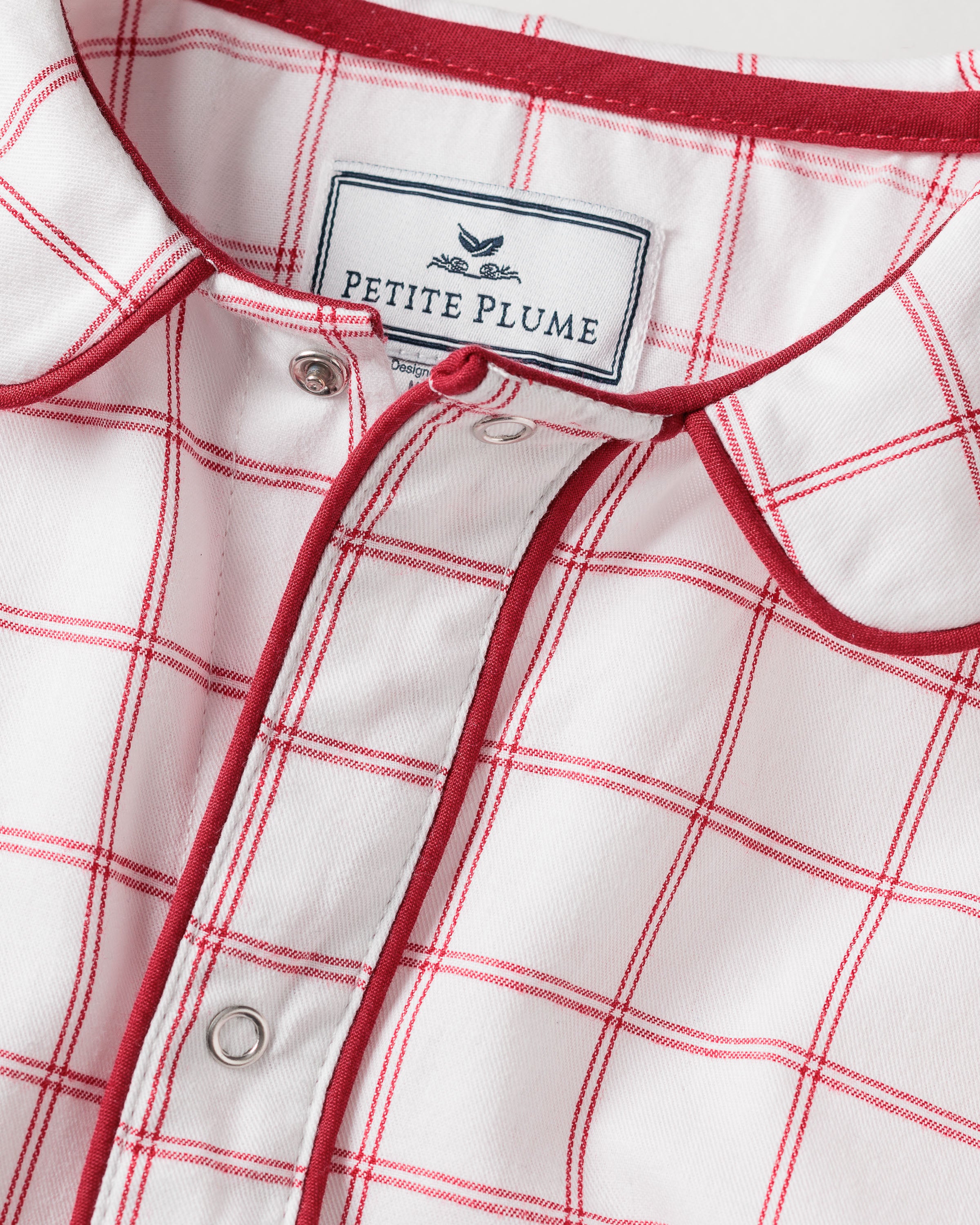 Close-up of a Babys Twill Cambridge Romper in Garnet Tattersall by Petite Plume, showcasing a garnet-red and white checkered pattern. The romper features a collar with red piping and is partially buttoned. Made from flame-retardant fabric for safety and style.