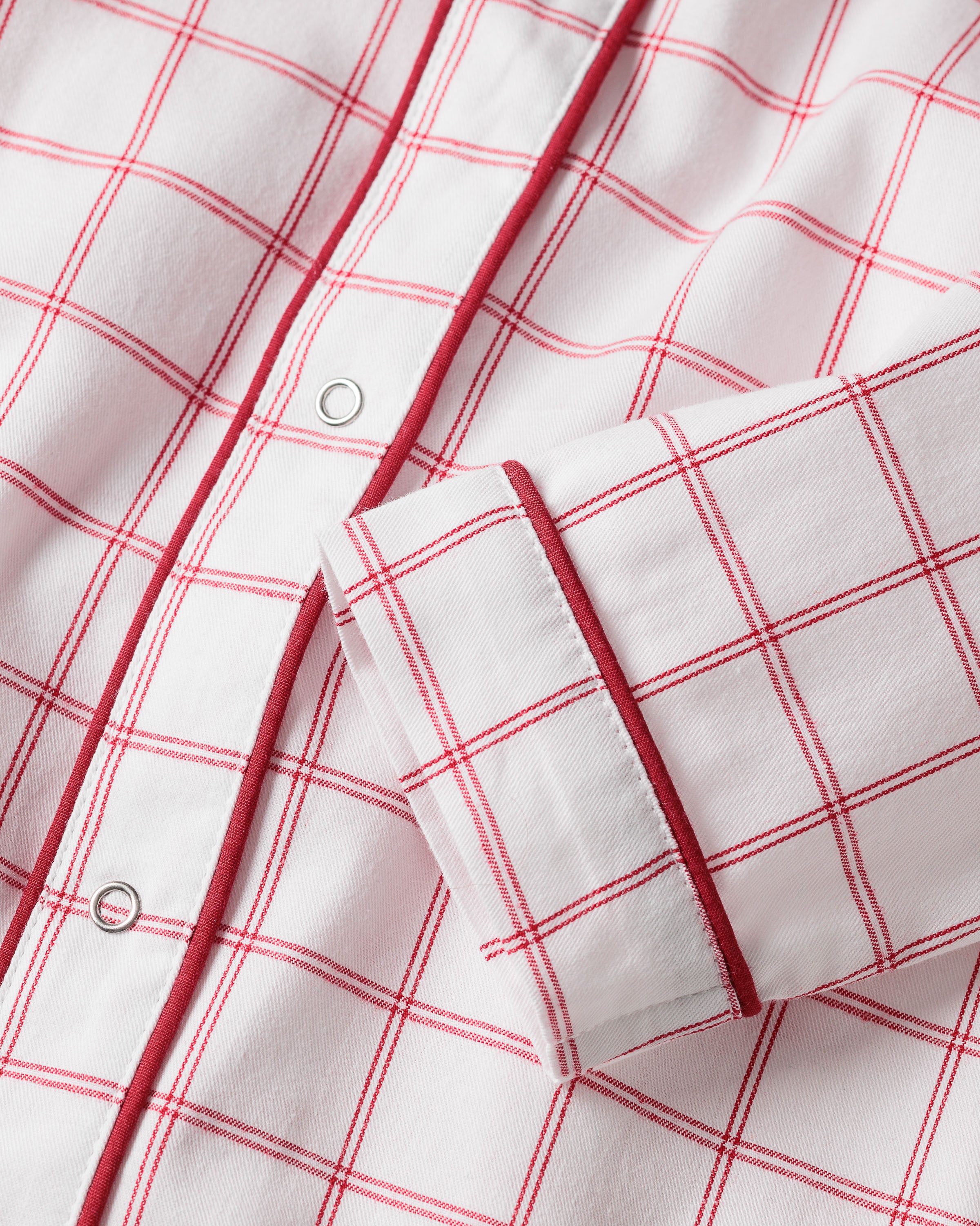 A close-up of the Babys Twill Cambridge Romper in Garnet Tattersall by Petite Plume shows its garnet plaid pattern. Ideal for childrens sleepwear, the image highlights a white shirt with red piping on the snap-button placket and sleeve cuff.