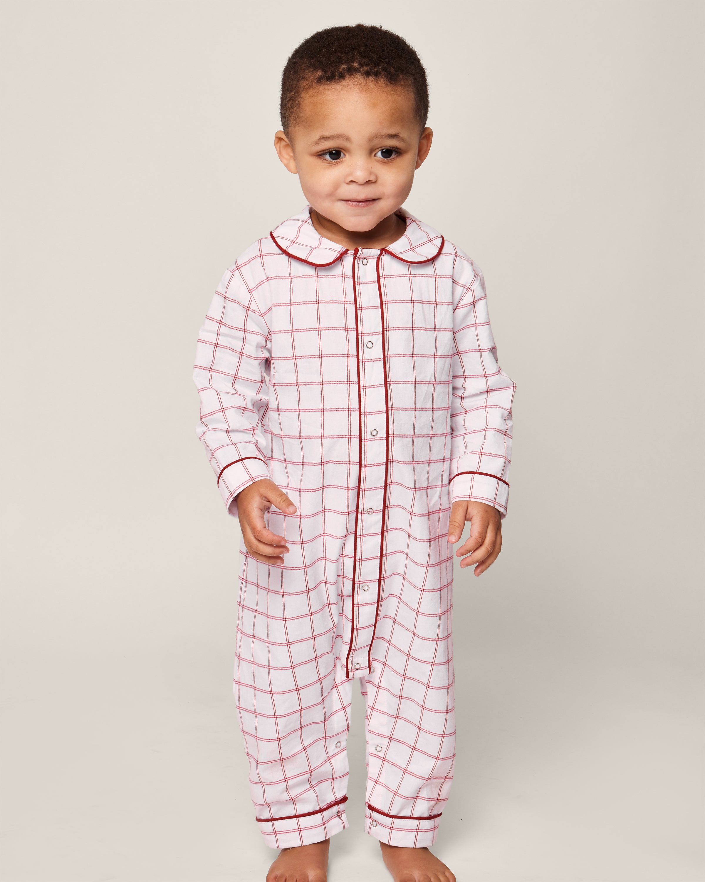A toddler, ready for bedtime in safe childrens sleepwear, stands against a plain background wearing the Petite Plume Babys Twill Cambridge Romper in Garnet Tattersall. The romper has a button-up front and Peter Pan collar and is made from flame-retardant fabric.