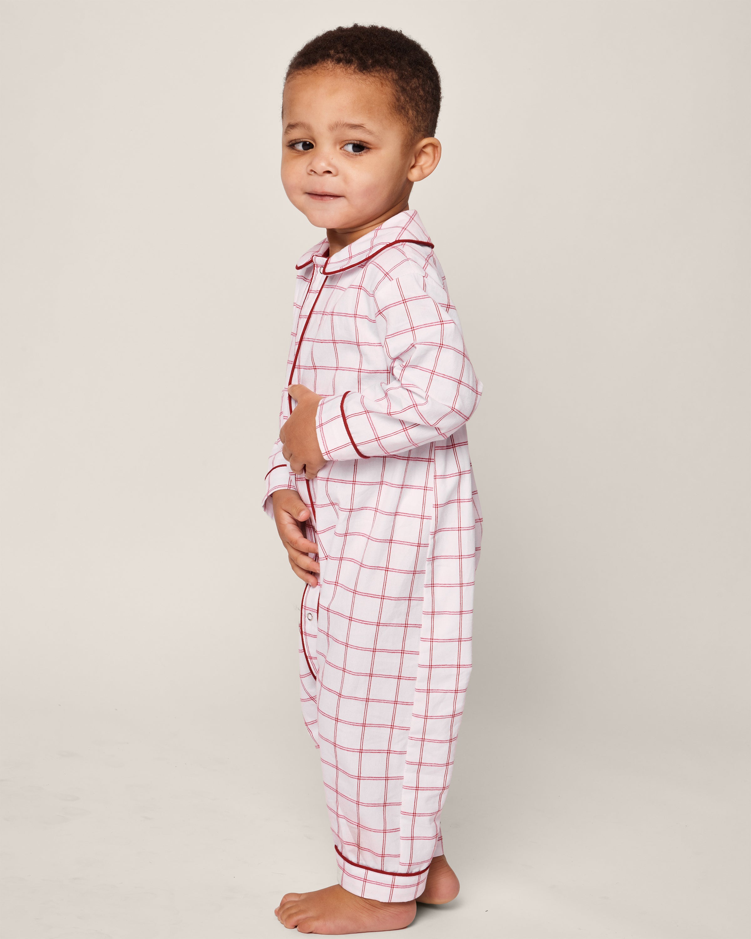 A young child stands sideways, smiling in a Petite Plume Babys Twill Cambridge Romper in Garnet Tattersall. The flame-retardant design ensures safety and comfort. The neutral background highlights the childs playful and relaxed demeanor.