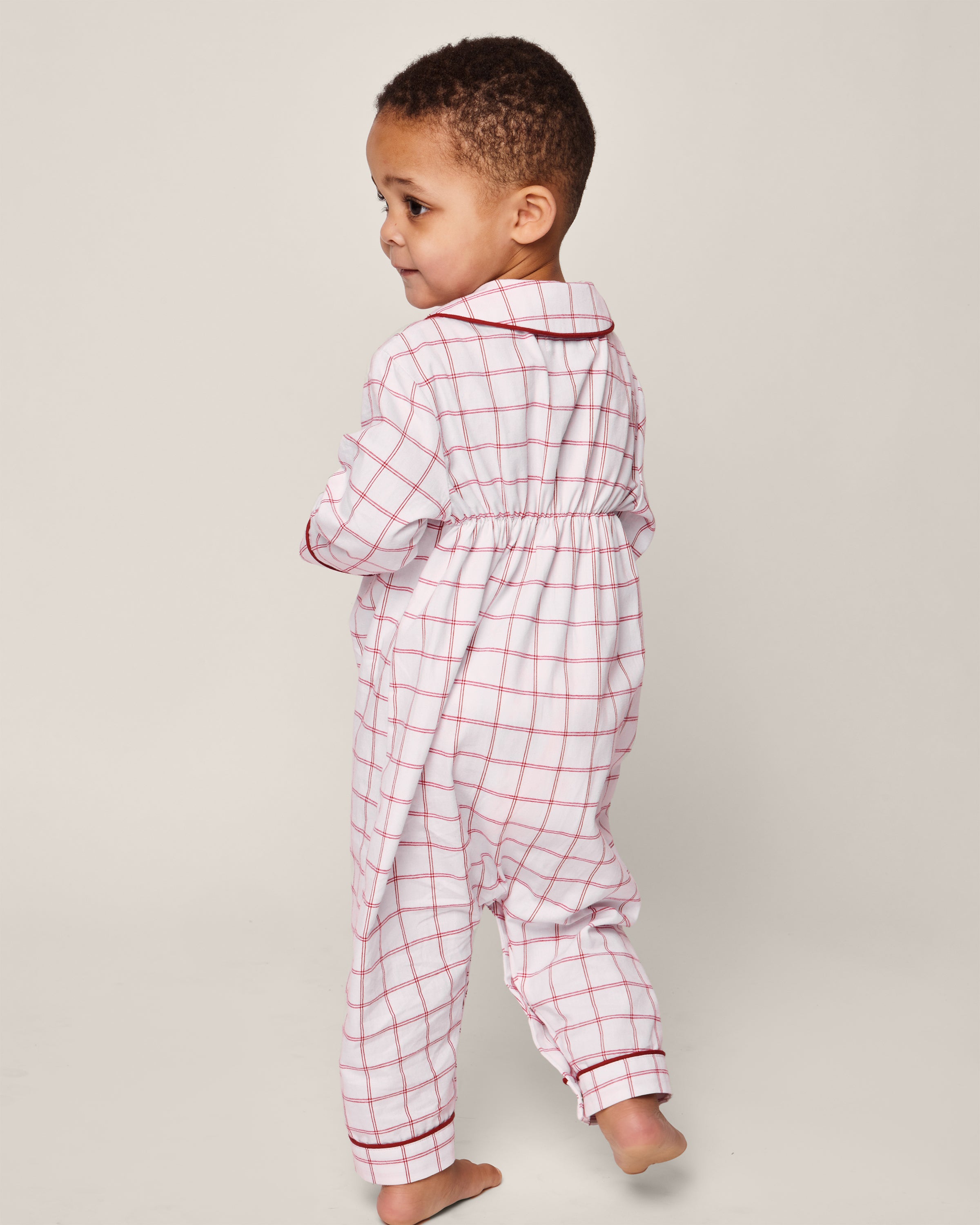 A young child in a Petite Plume Babys Twill Cambridge Romper in Garnet Tattersall is turned slightly away, looking right. This childrens onesie features a collar, an elastic waist, and flame-retardant fabric. The background is plain and light-colored.