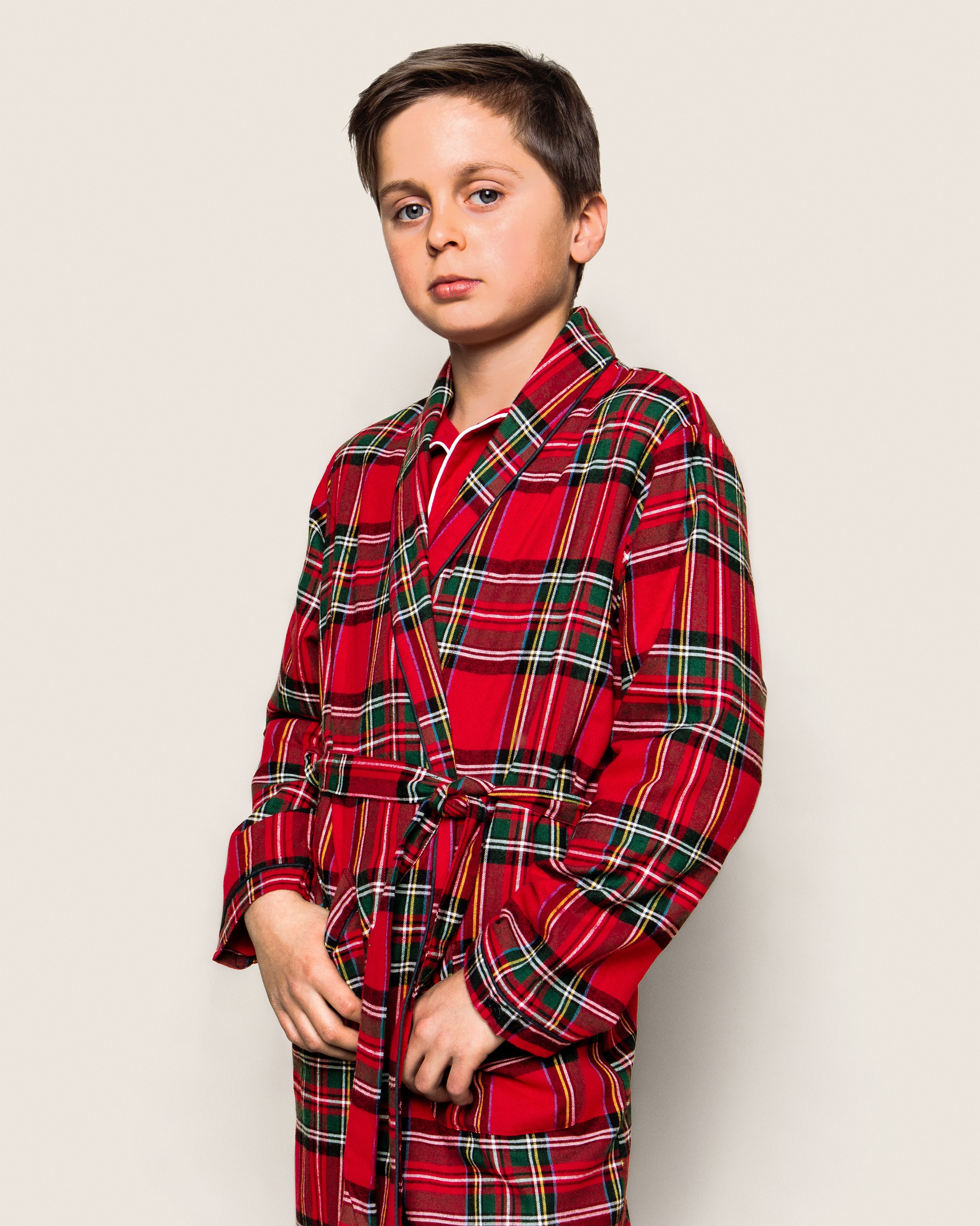 A young boy stands against a plain backdrop, wearing Petite Plumes Kids Brushed Cotton Robe in Imperial Tartan. He looks at the camera with a neutral expression, holding the yarn-dyed cotton belt with both hands.